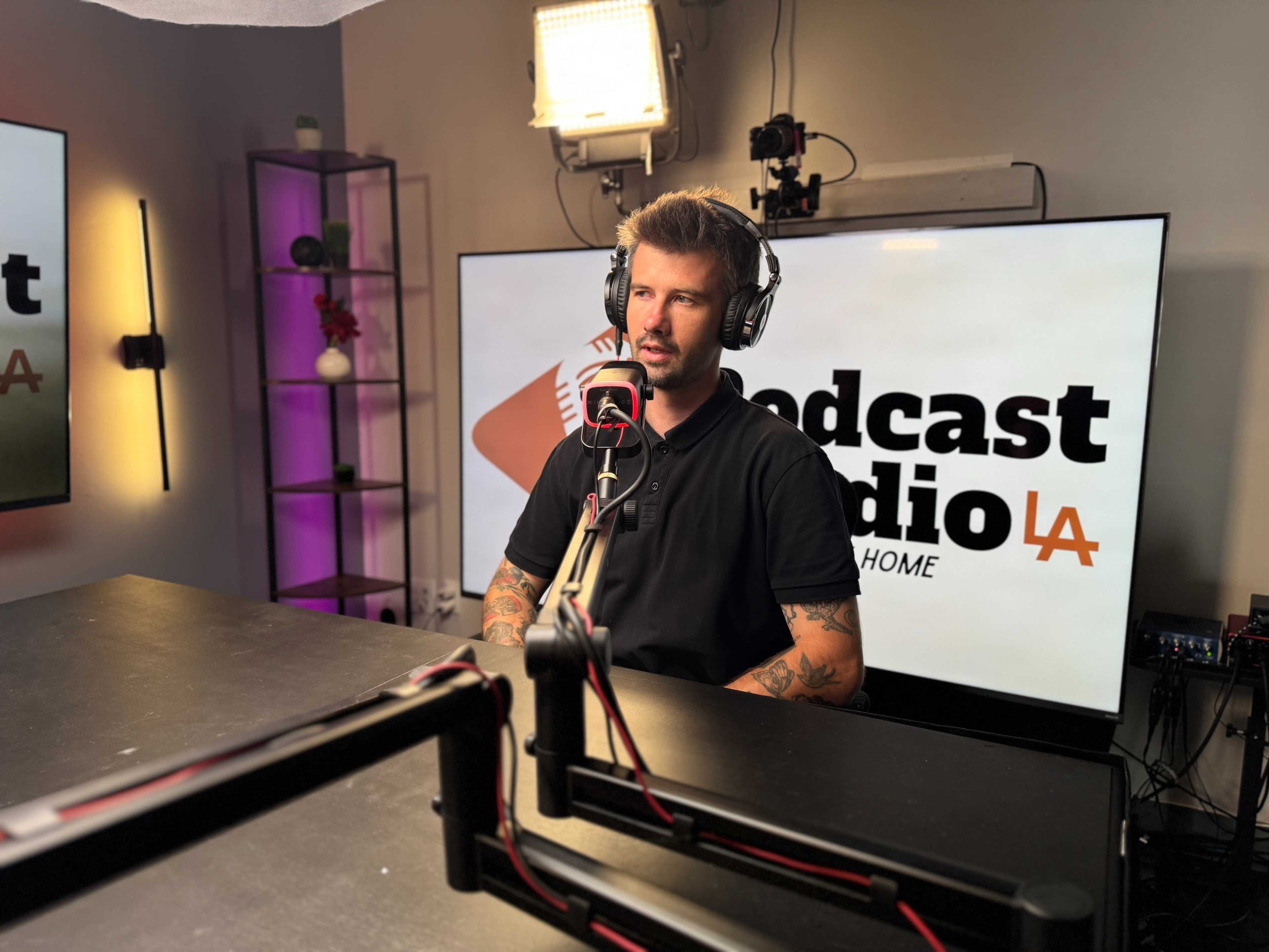 A man wearing headphones sits at a podcast studio desk in front of a large screen displaying the words "Podcast Studio LA."