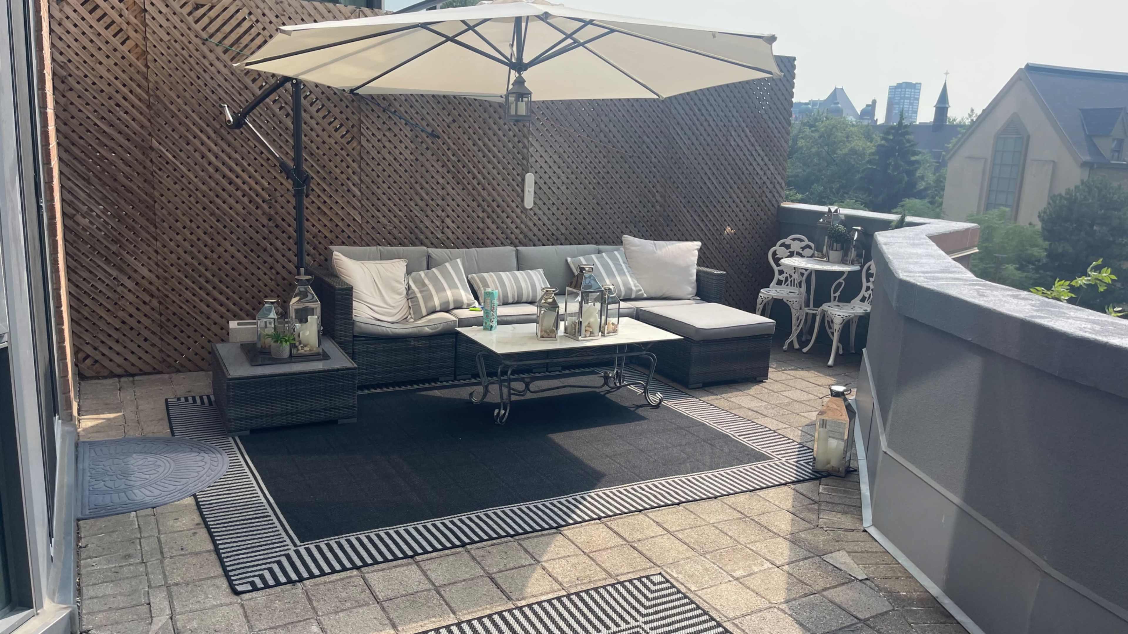 The image shows a rooftop terrace with a sectional sofa, a large umbrella, and a coffee table surrounded by decorative elements and cityscape in the background.