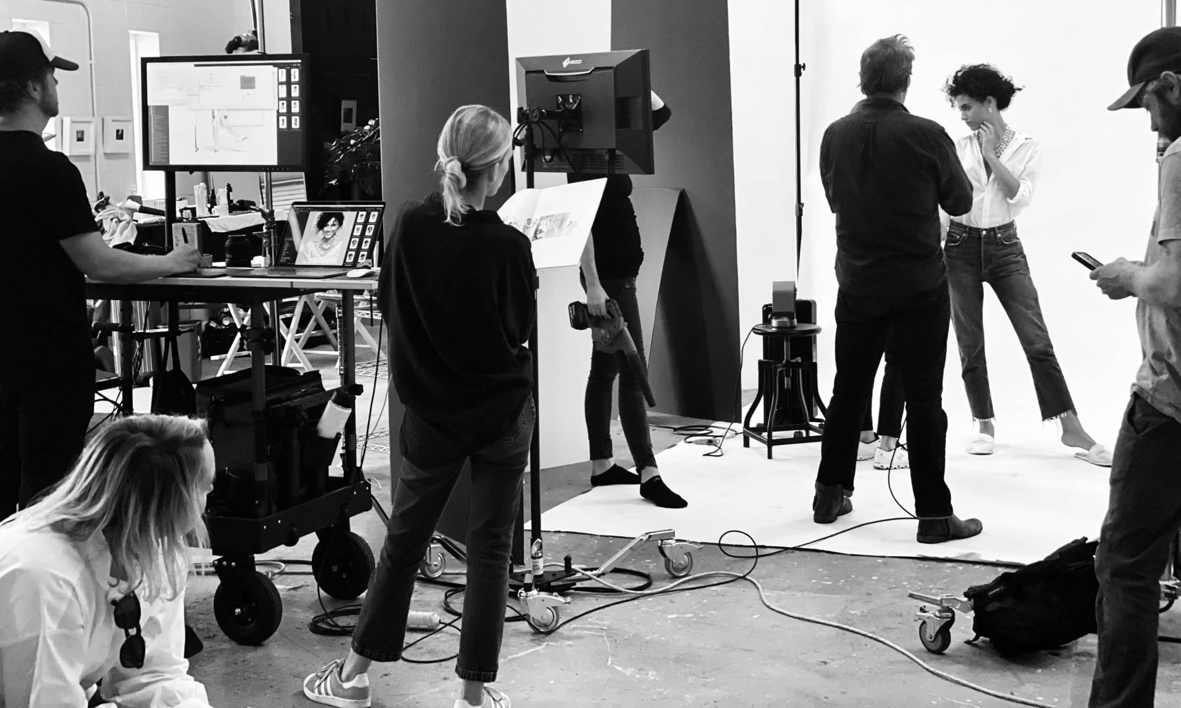 A group of people is working on a photoshoot in a studio, with a model posing in front of a camera while others assist and observe.