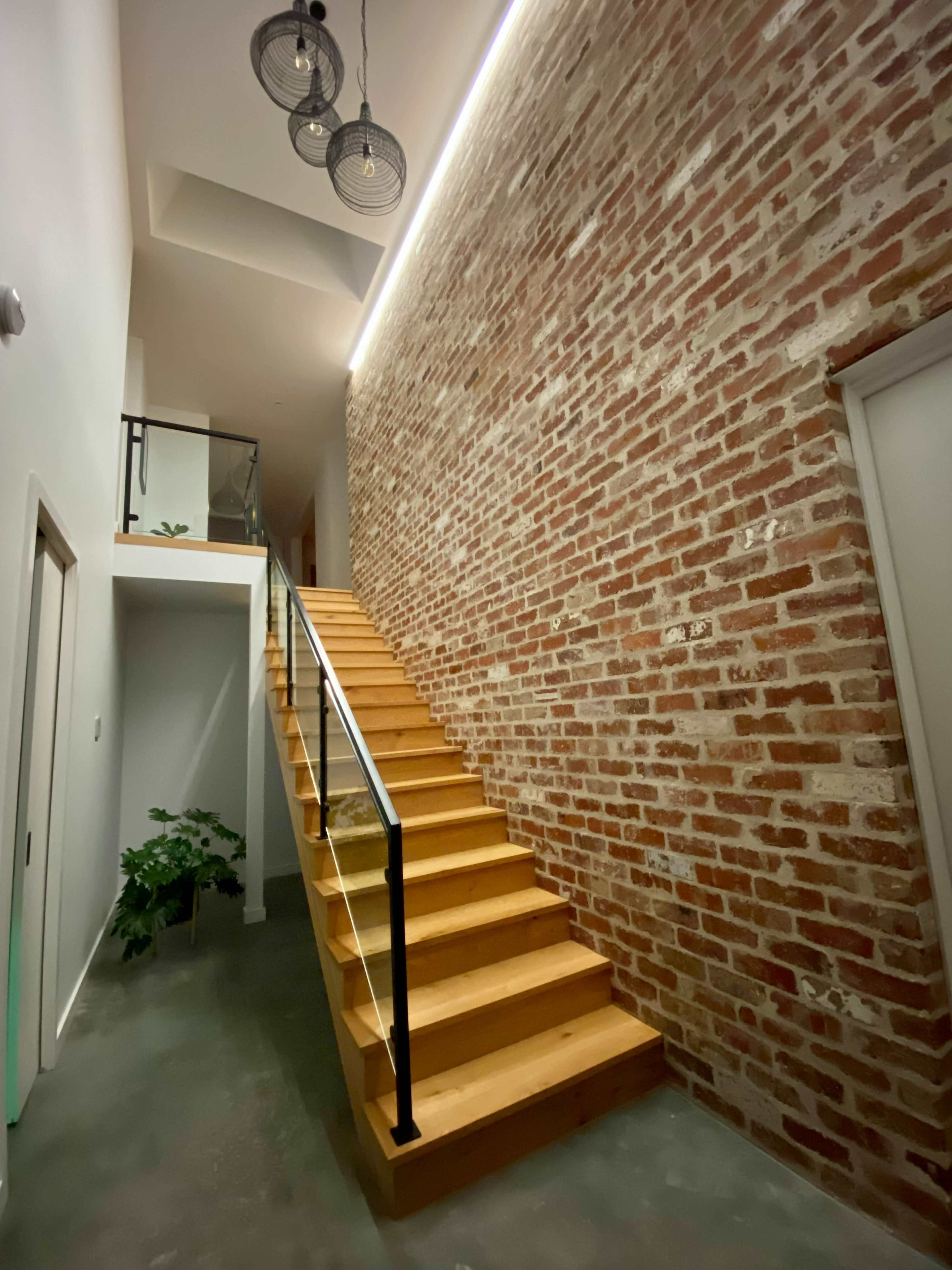 A wooden stairway leading to an upper level, flanked by a brick wall and illuminated by pendant lights and recessed lighting.