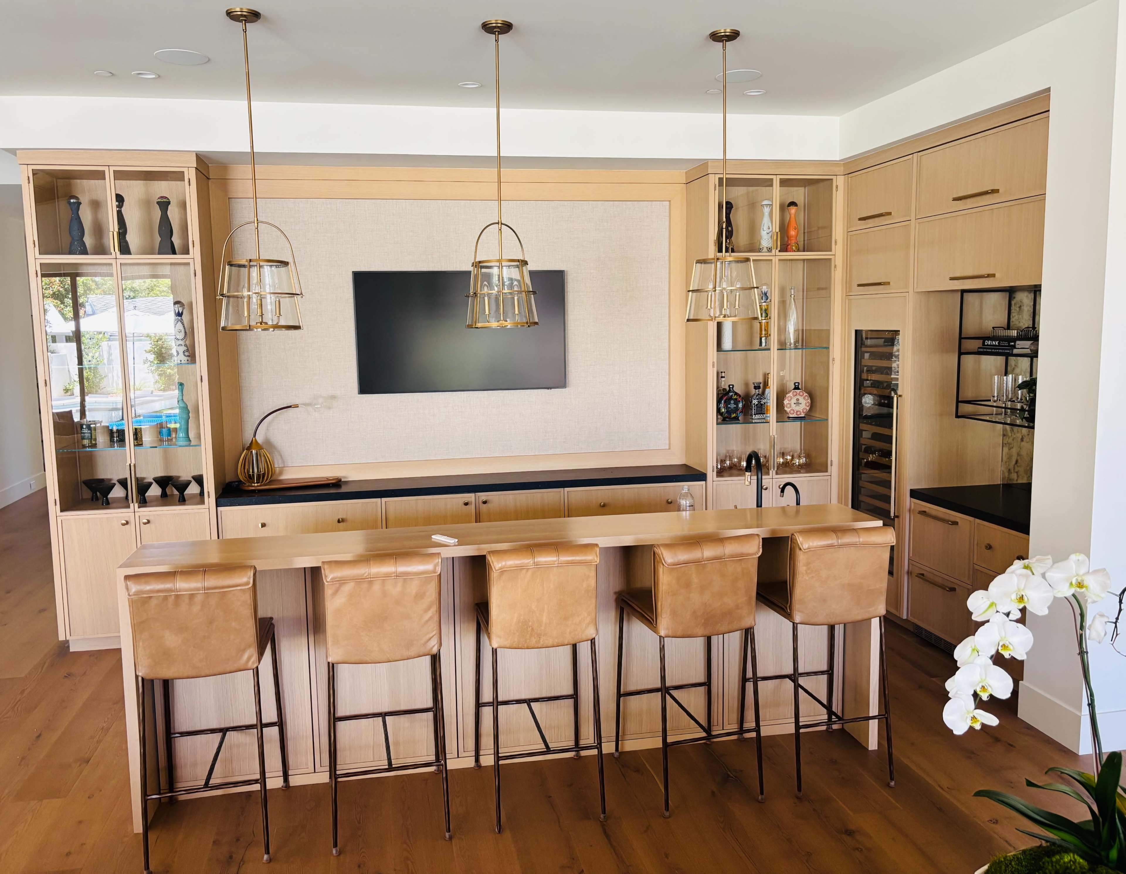 The image shows a modern home bar area with a wooden counter, high stools, and decorative shelves featuring various drinks and glassware.