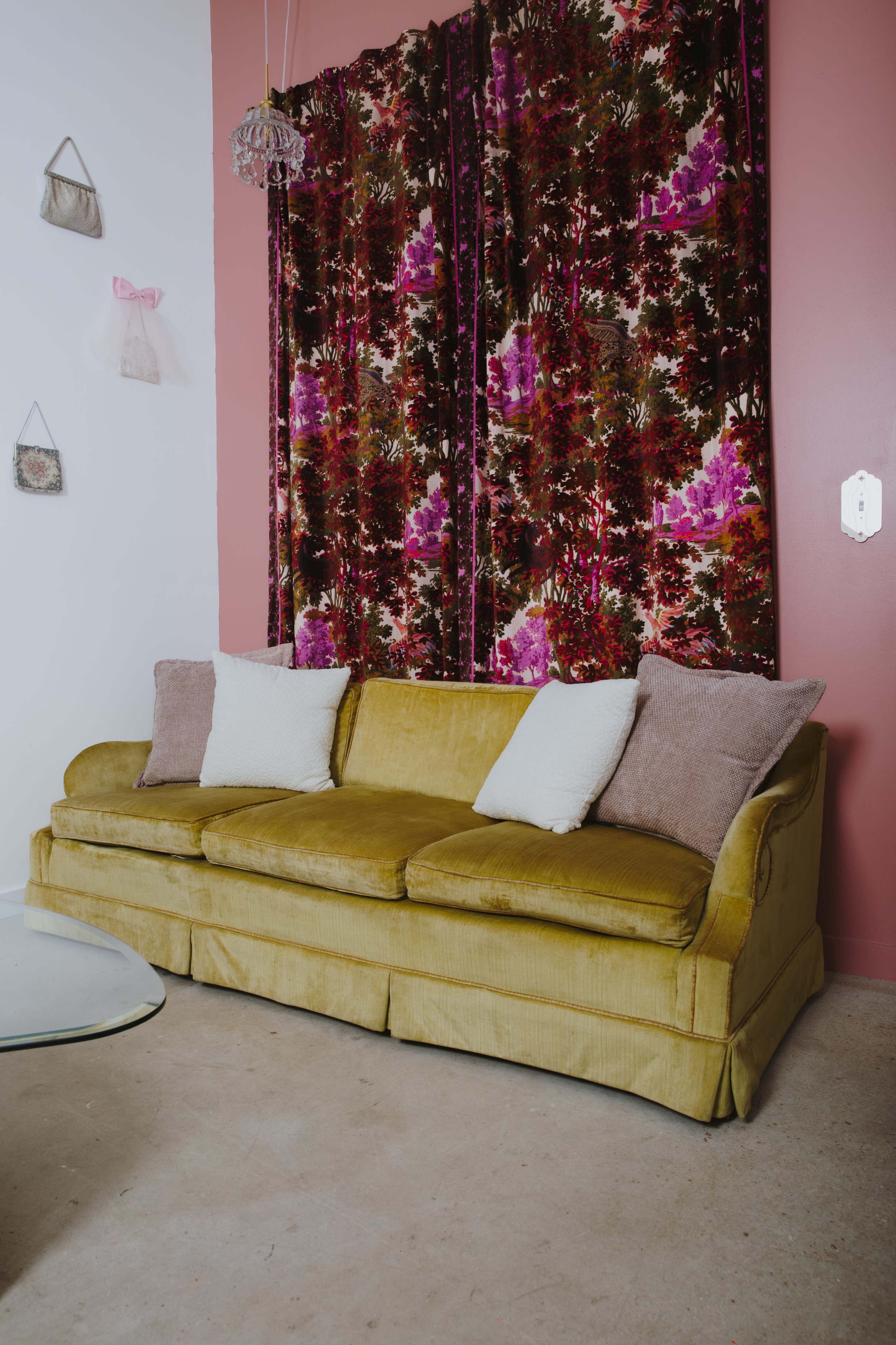 A yellow sofa with two white and two pink cushions is positioned in front of a patterned curtain in a room with a pink wall and a glass coffee table.