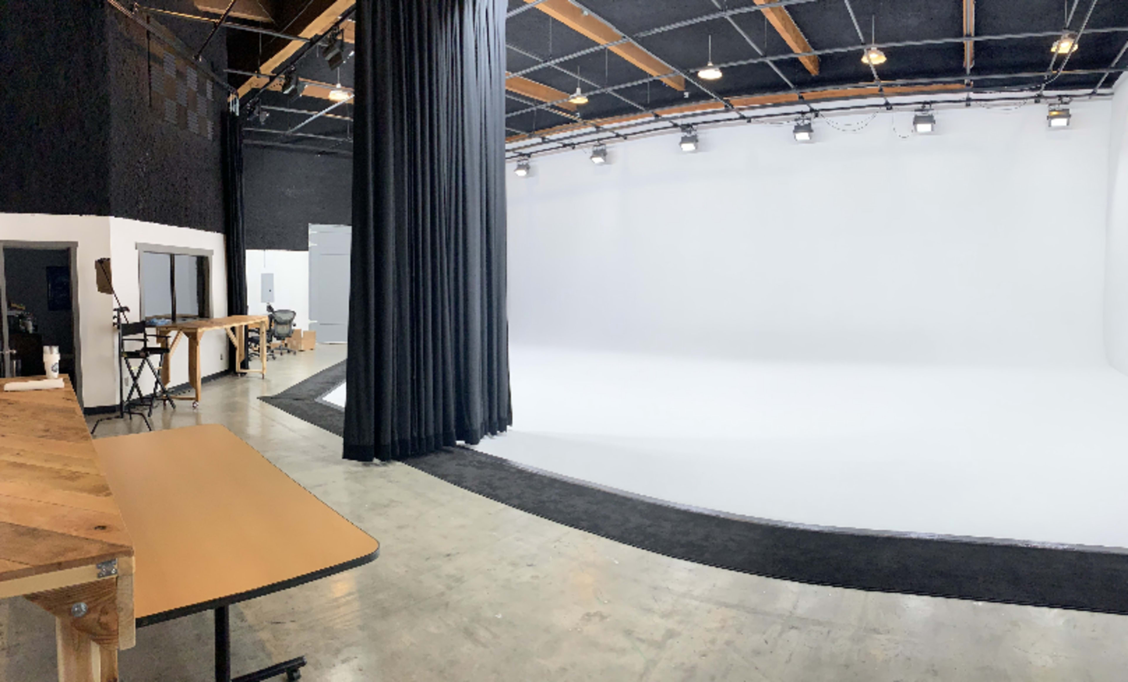 The image shows a spacious studio with a white backdrop, black curtains, and a wooden table in the foreground.
