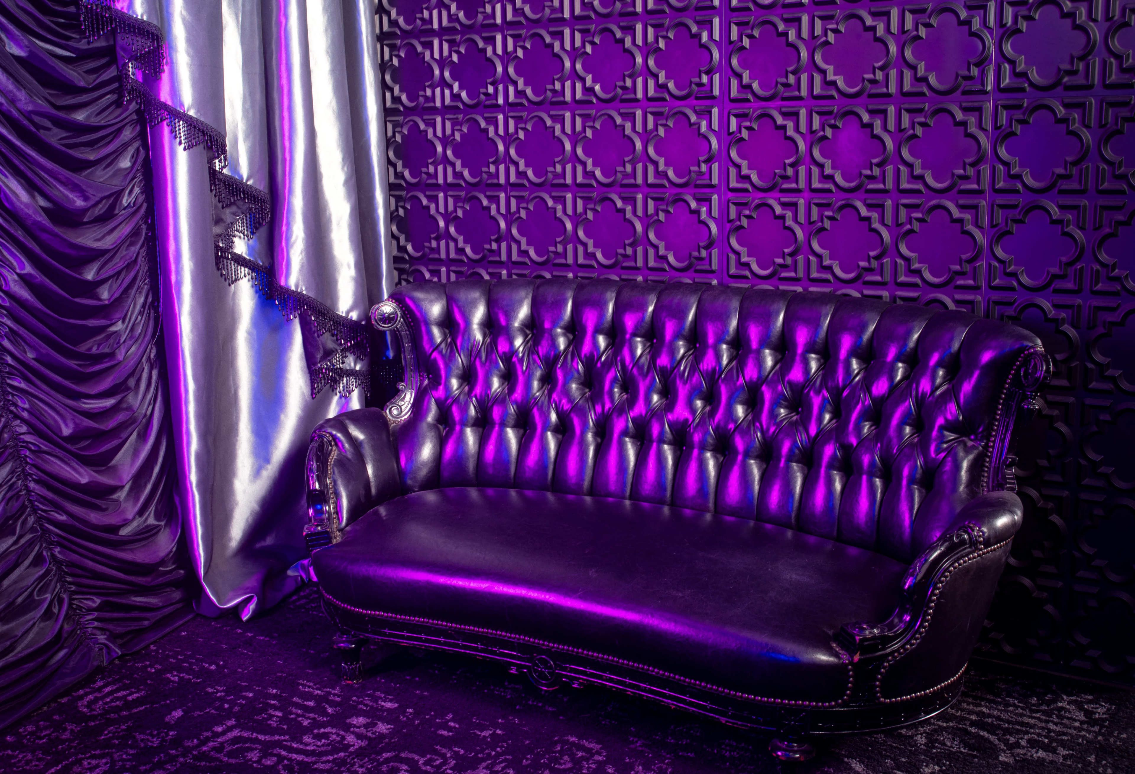 A black leather tufted sofa set against a patterned purple and black wall, with draped silver curtains nearby.