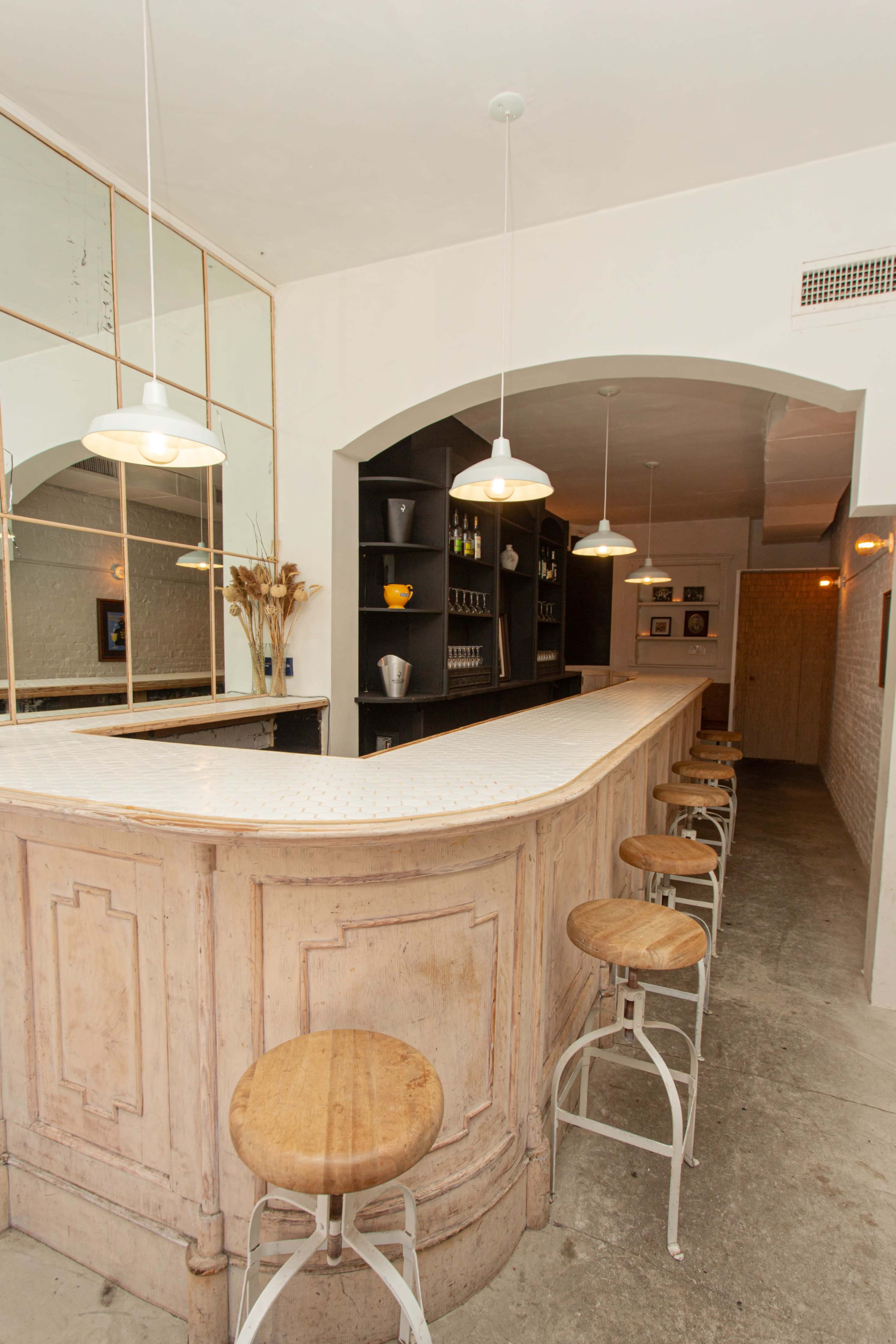 The image shows a bar area with a curved countertop, several stools, and shelves displaying bottles, illuminated by hanging lights.