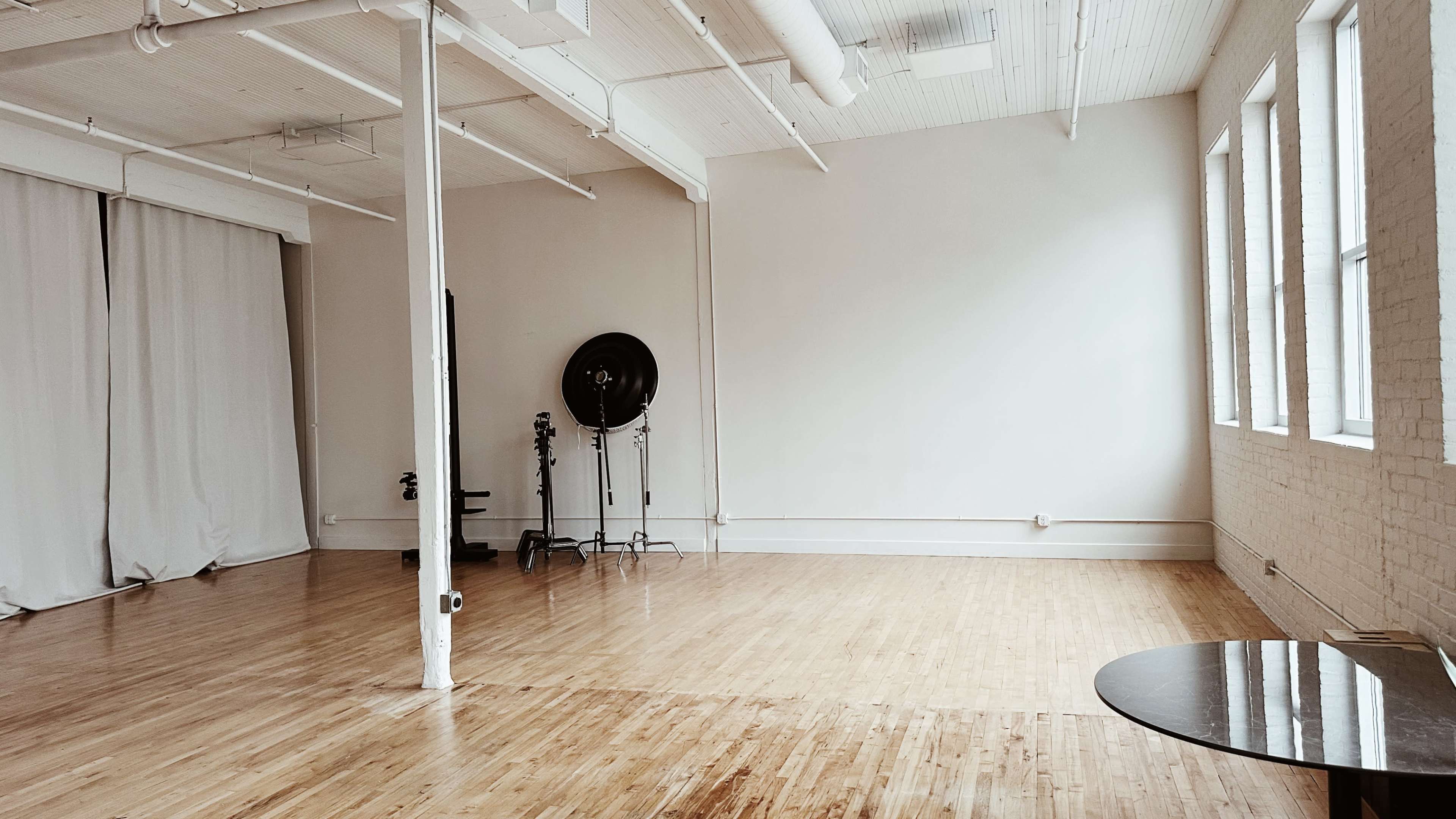 A spacious, empty room with hardwood floors, large windows, and a few pieces of studio equipment against one wall.