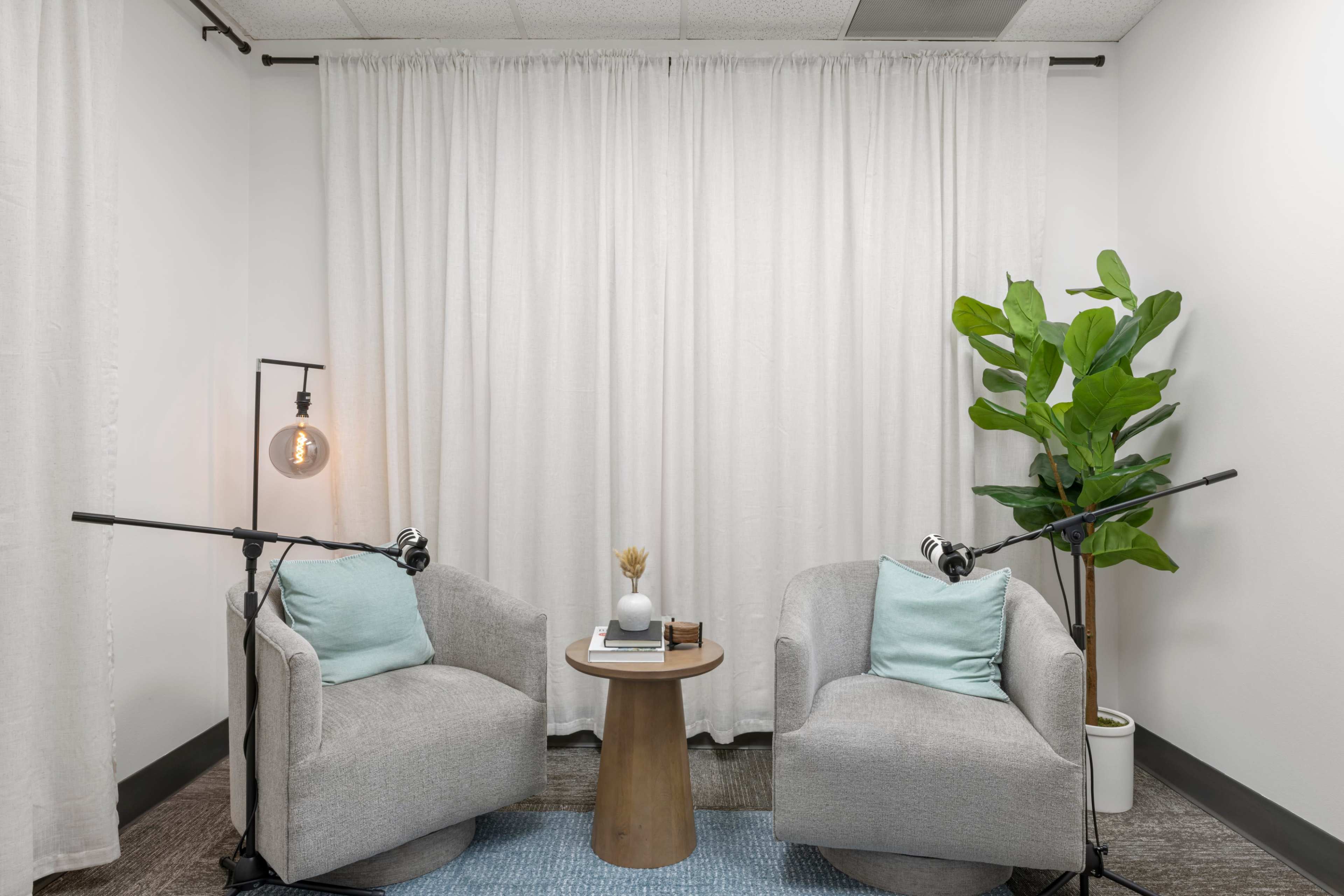The image shows a small, modern space with two gray armchairs, a round wooden table, and a potted plant, all arranged in front of a white curtain.