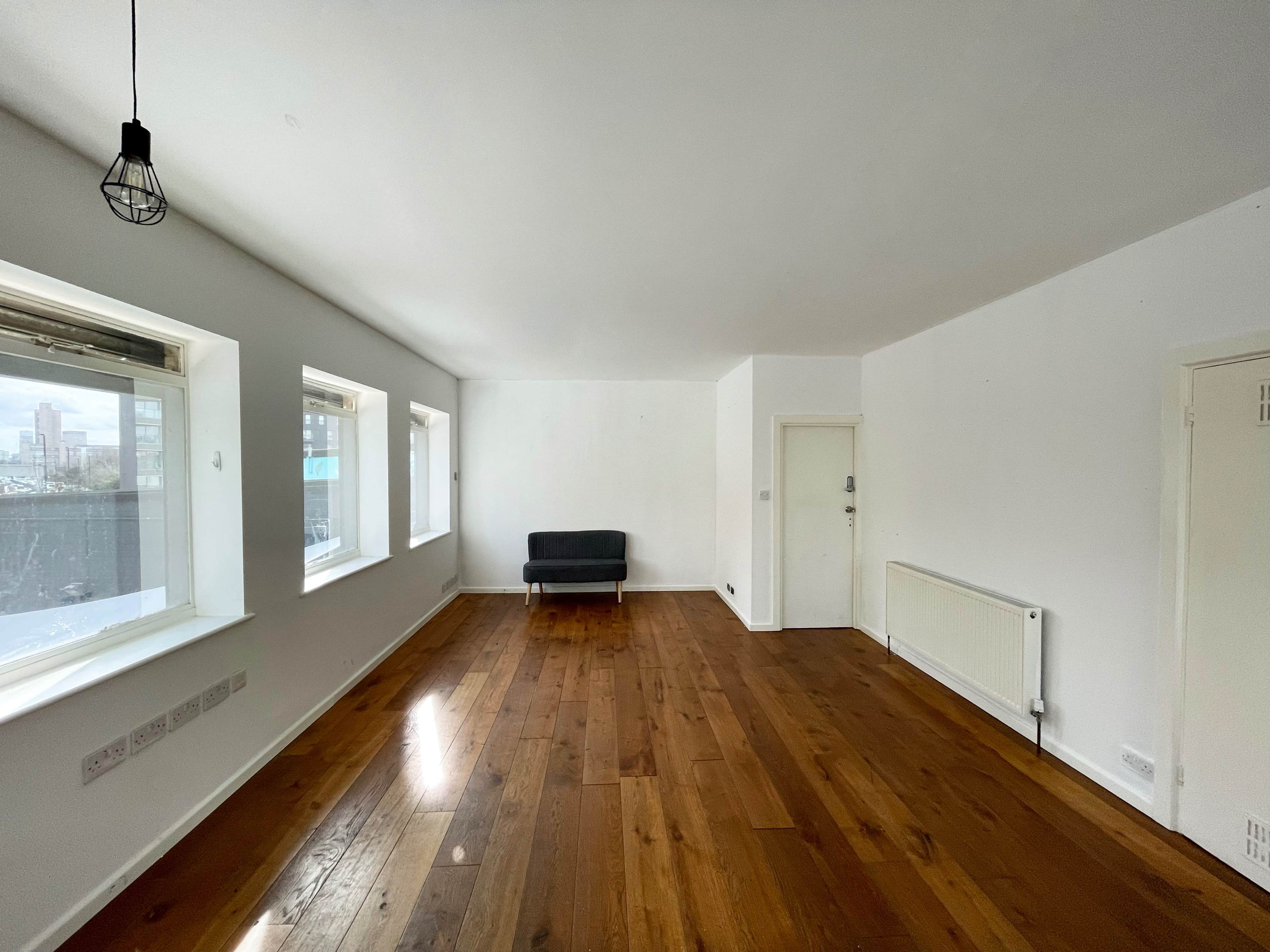 A spacious, empty room with wooden flooring, a single gray sofa, large windows, and a door leading to another area.