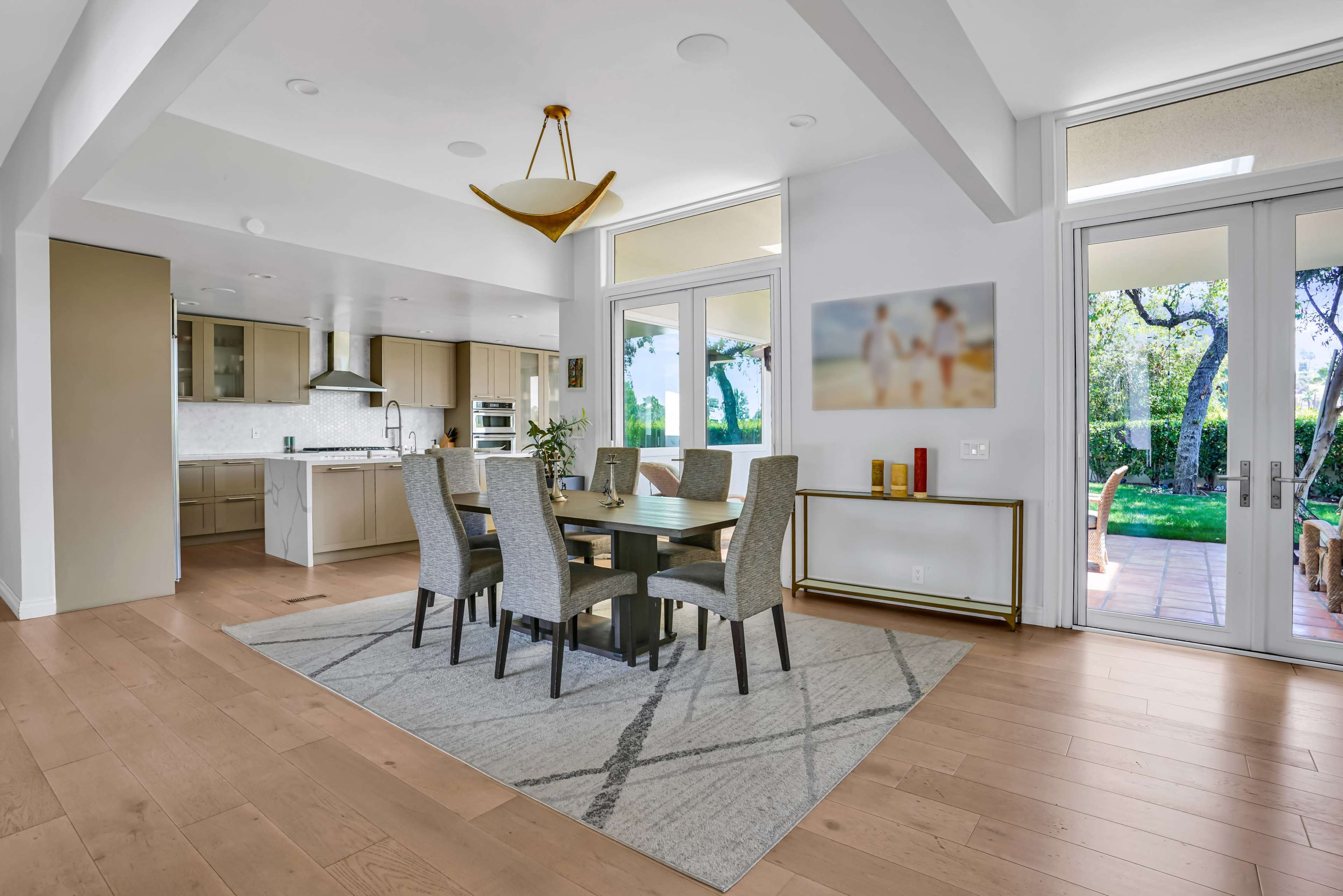 A modern dining area features a rectangular wooden table surrounded by light gray upholstered chairs, with large windows providing natural light and a view of the outdoors.