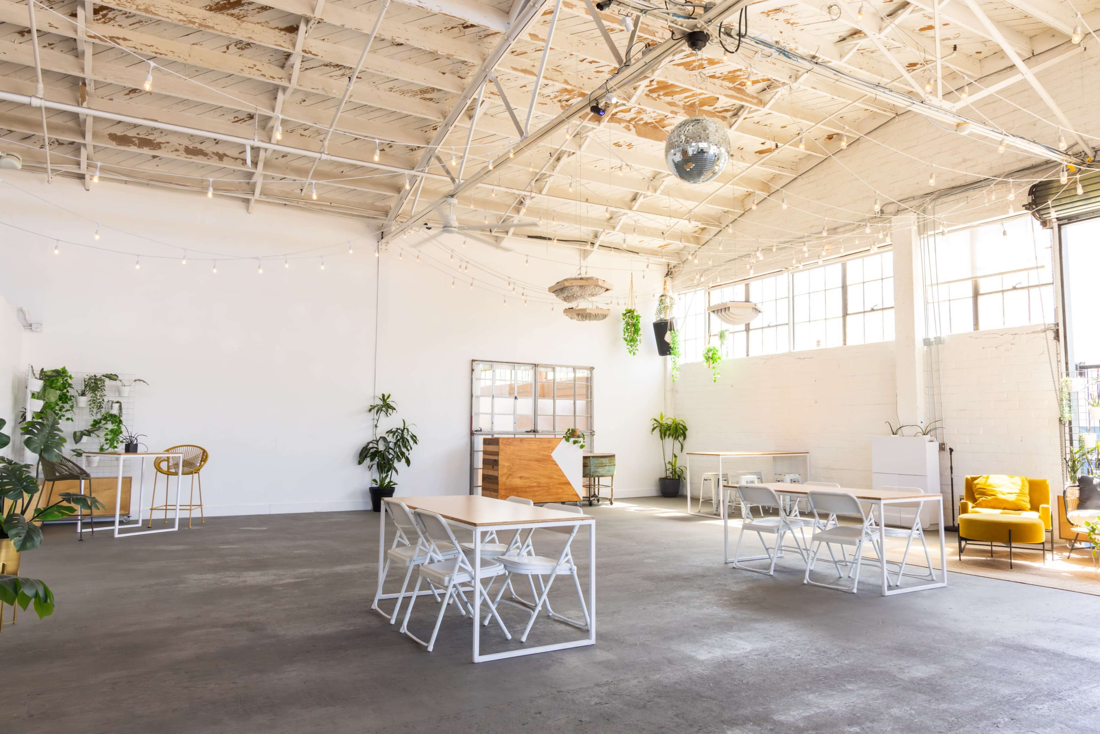 A spacious, well-lit industrial room features several white tables, chairs, and plants, with large windows allowing natural light to enter.