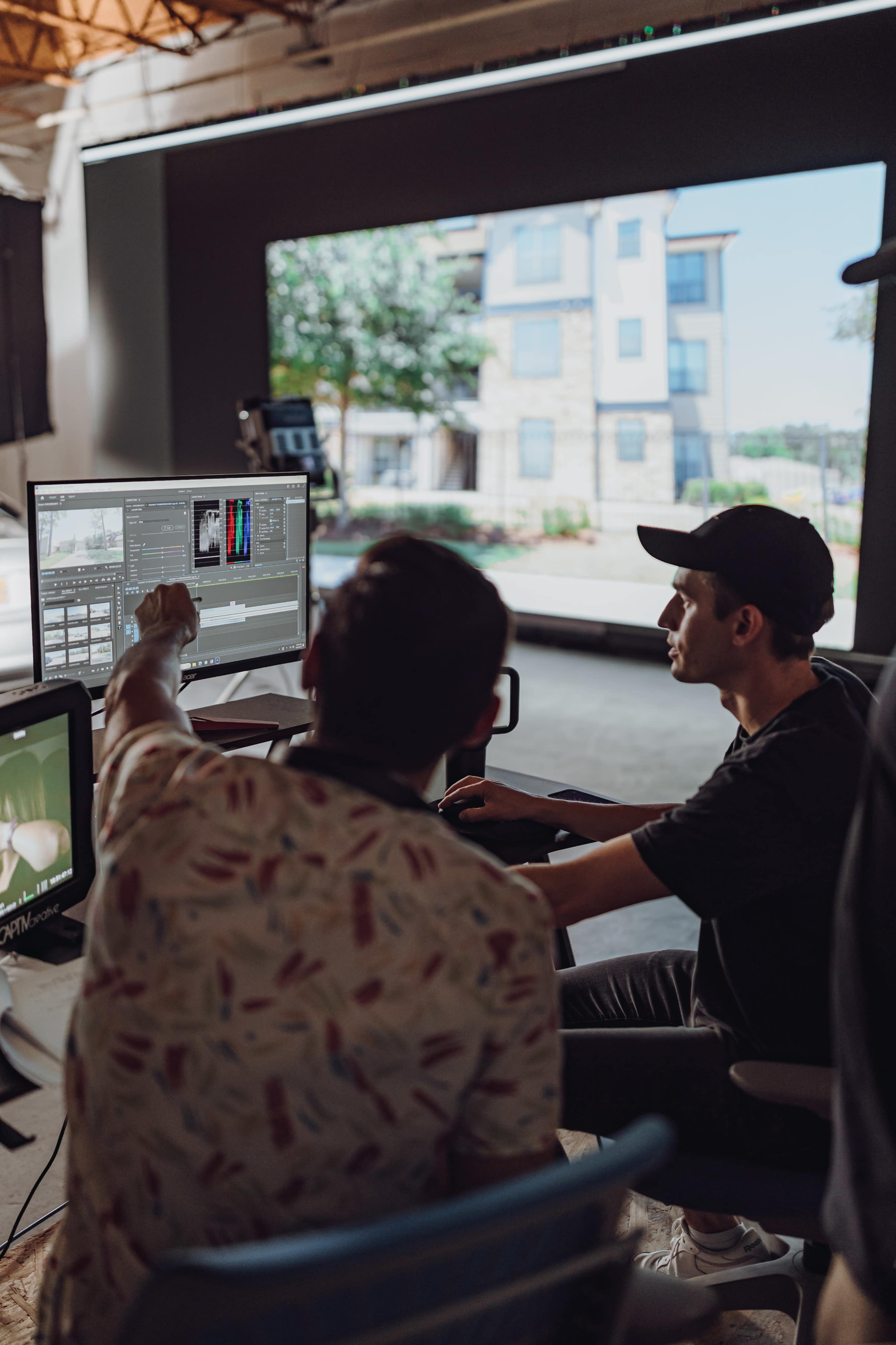 Two individuals are seated at a computer, reviewing video footage on multiple screens while discussing edits in a well-lit studio with large windows.