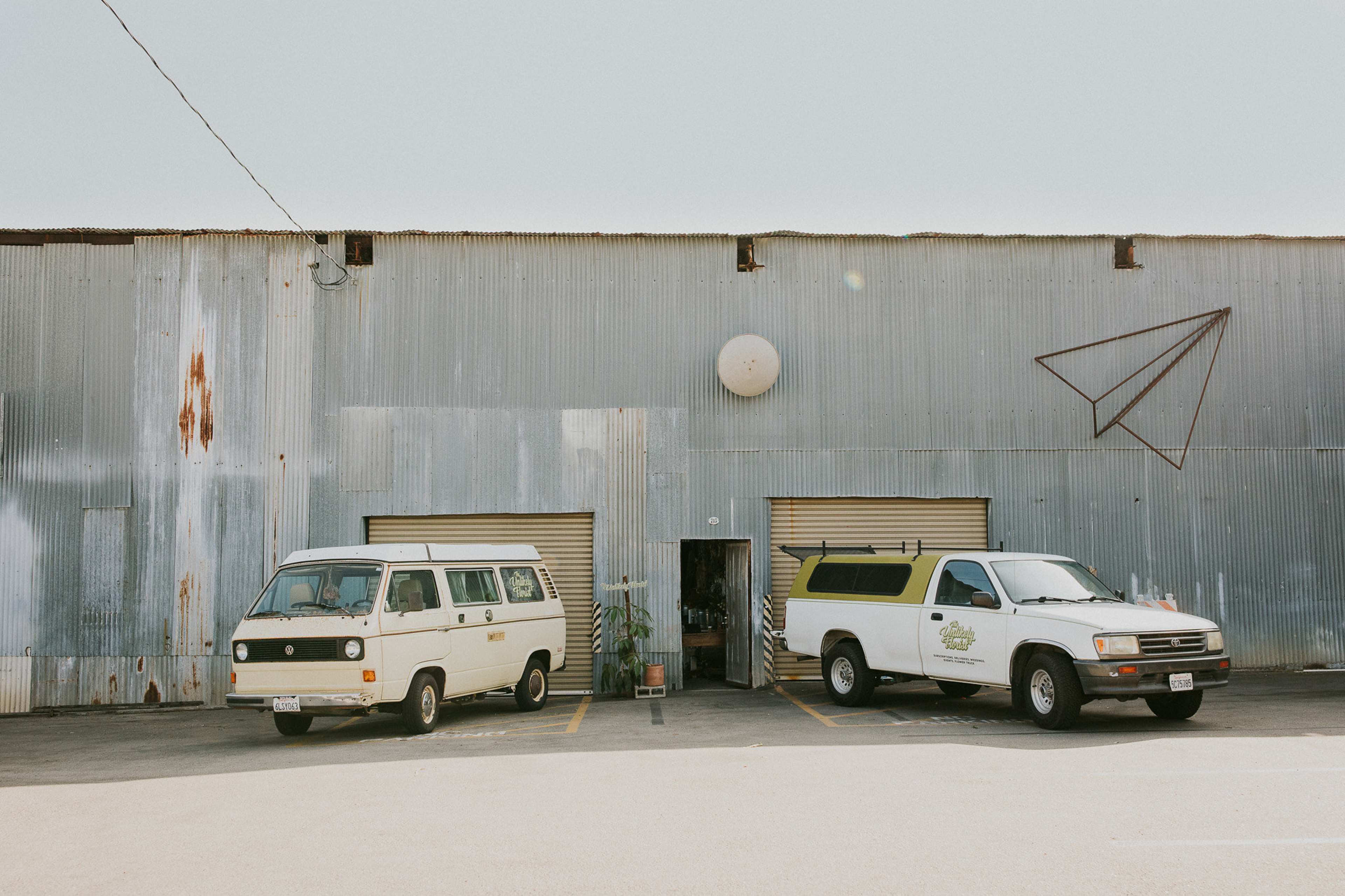A vintage van and a white pickup truck are parked outside a corrugated metal building with two roll-up doors.