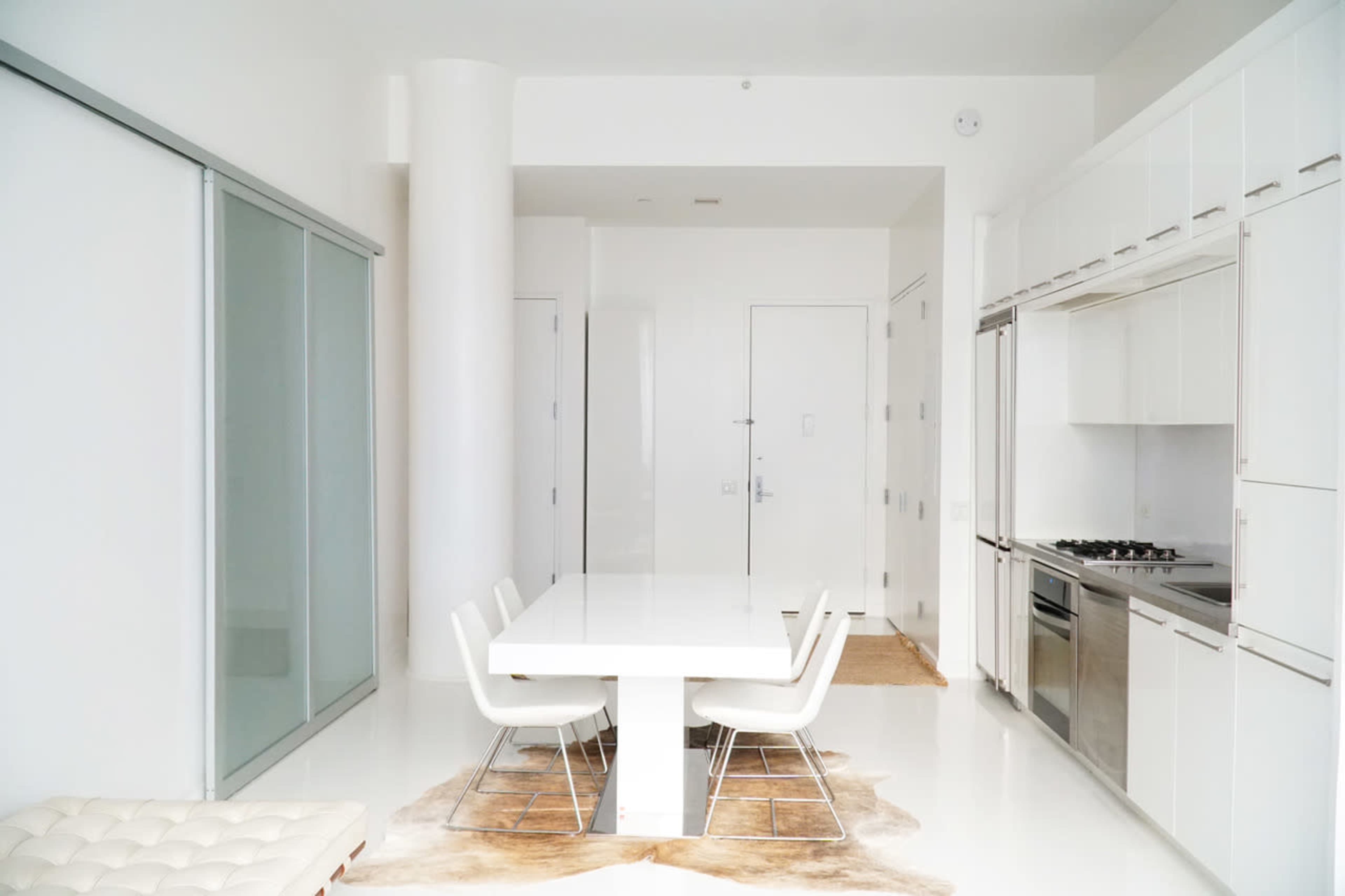 A modern kitchen features a sleek white dining table surrounded by four chairs, with minimalist cabinetry and a stainless steel stove against a bright, open space.