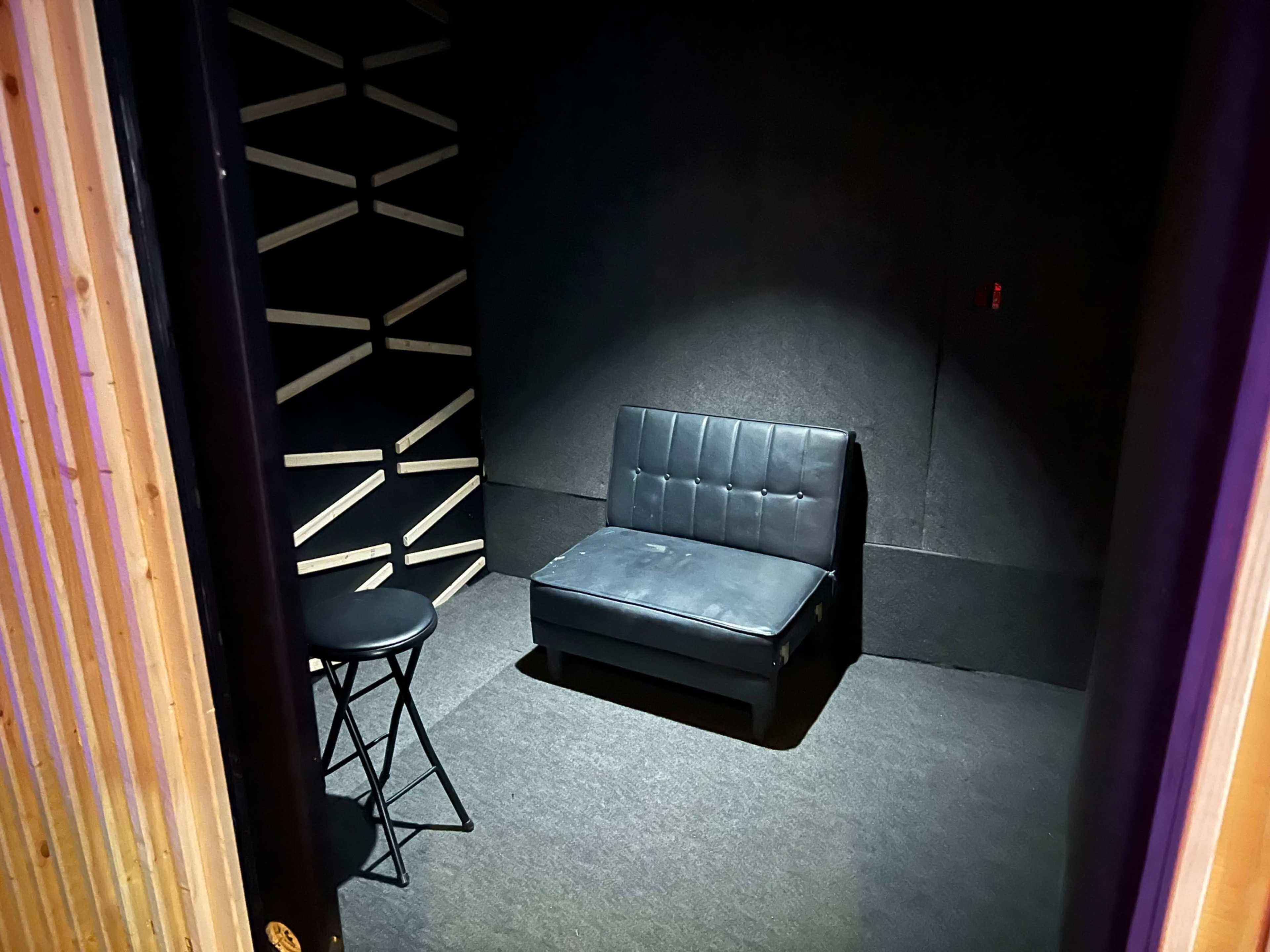 A small, dimly lit room featuring a black upholstered chair and a tall black stool against a textured wall.