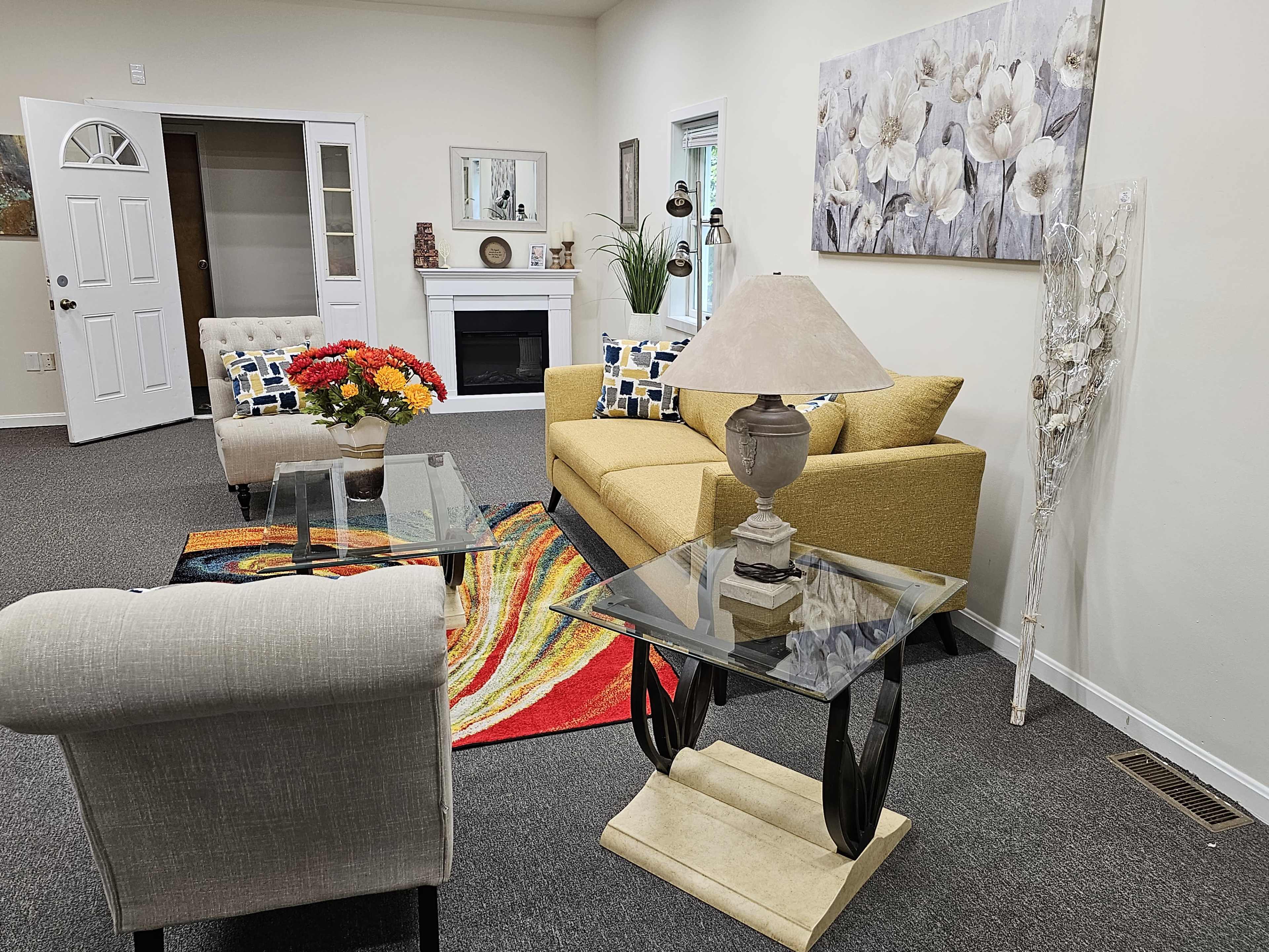 The image depicts a cozy living area with a yellow couch, glass tables, and a decorative lamp, all arranged on a patterned rug.