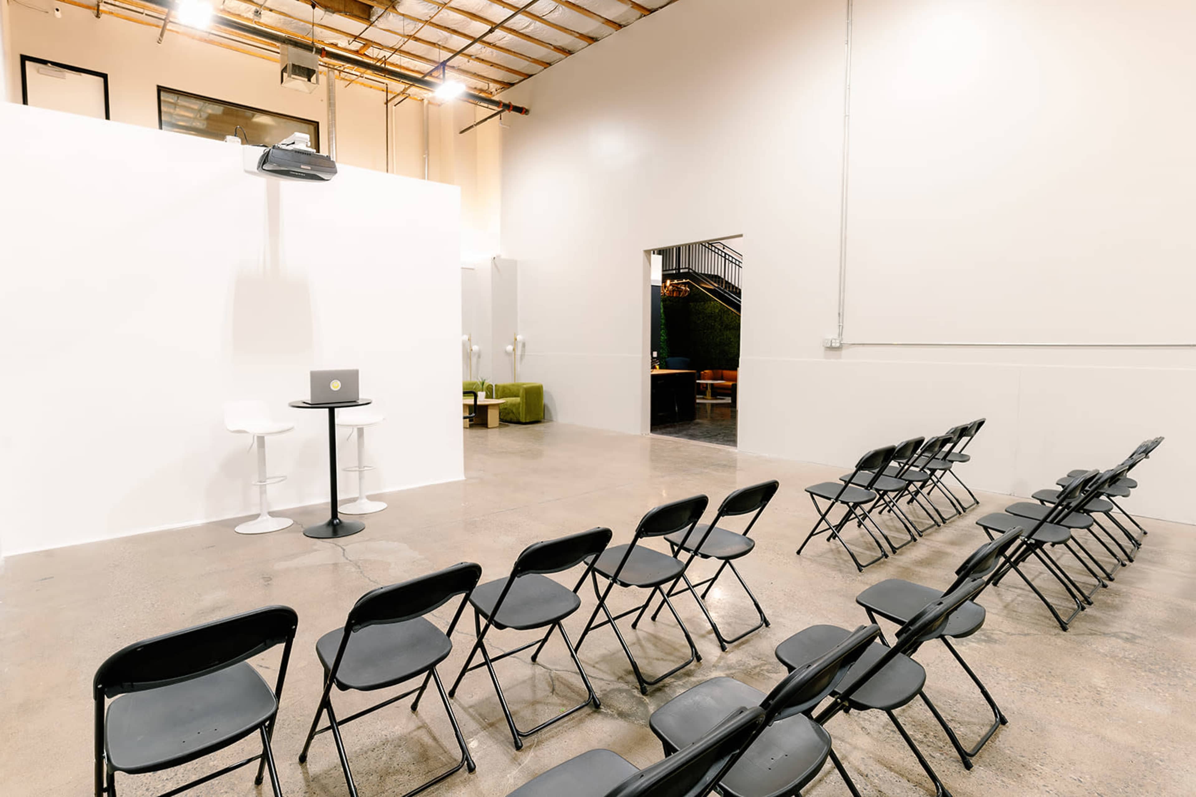 A spacious, minimalistic room with rows of black chairs facing a white partition and a small table with a laptop on it.