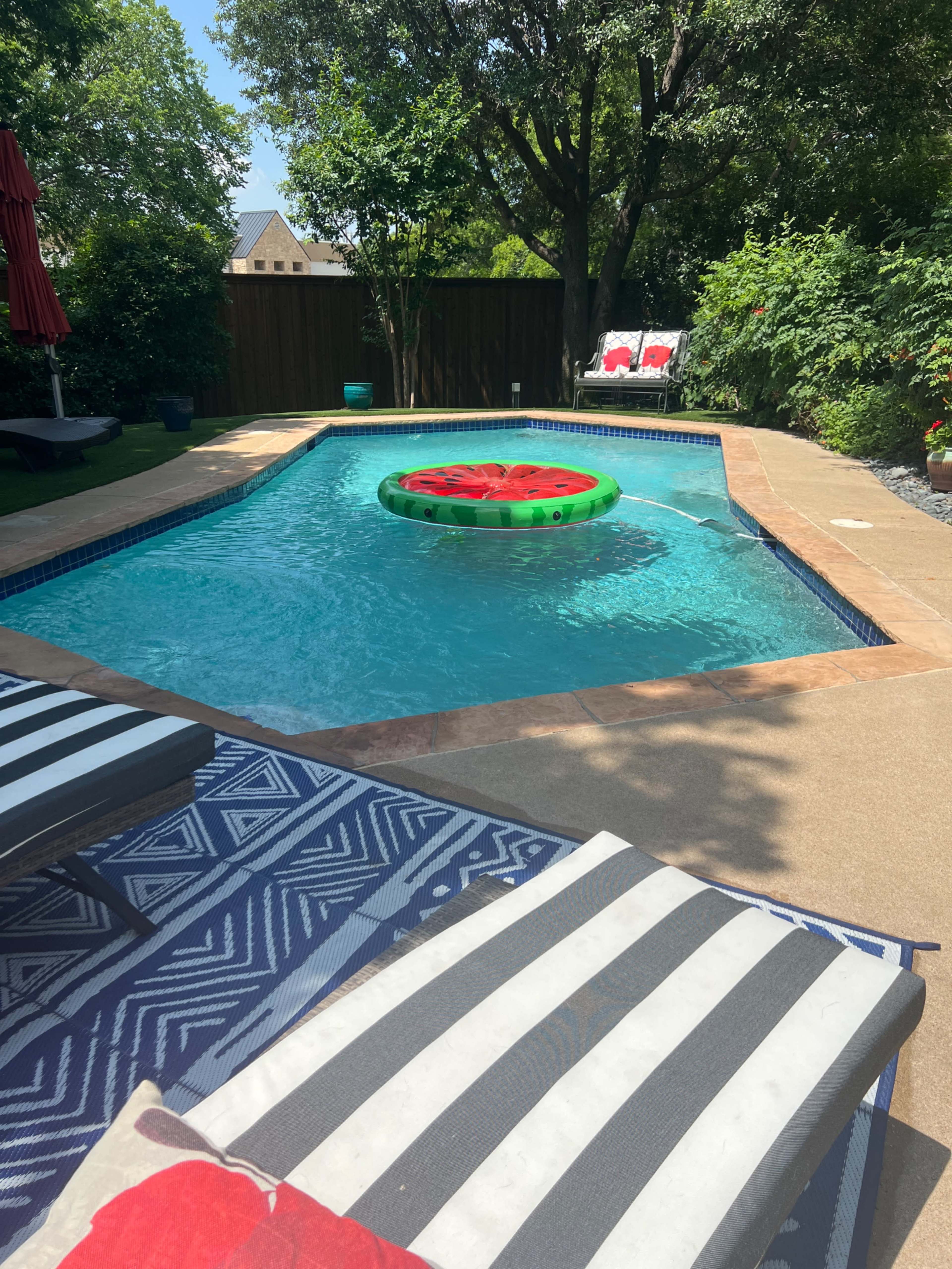 A green inflatable watermelon floats in a swimming pool surrounded by lounge chairs and lush greenery.