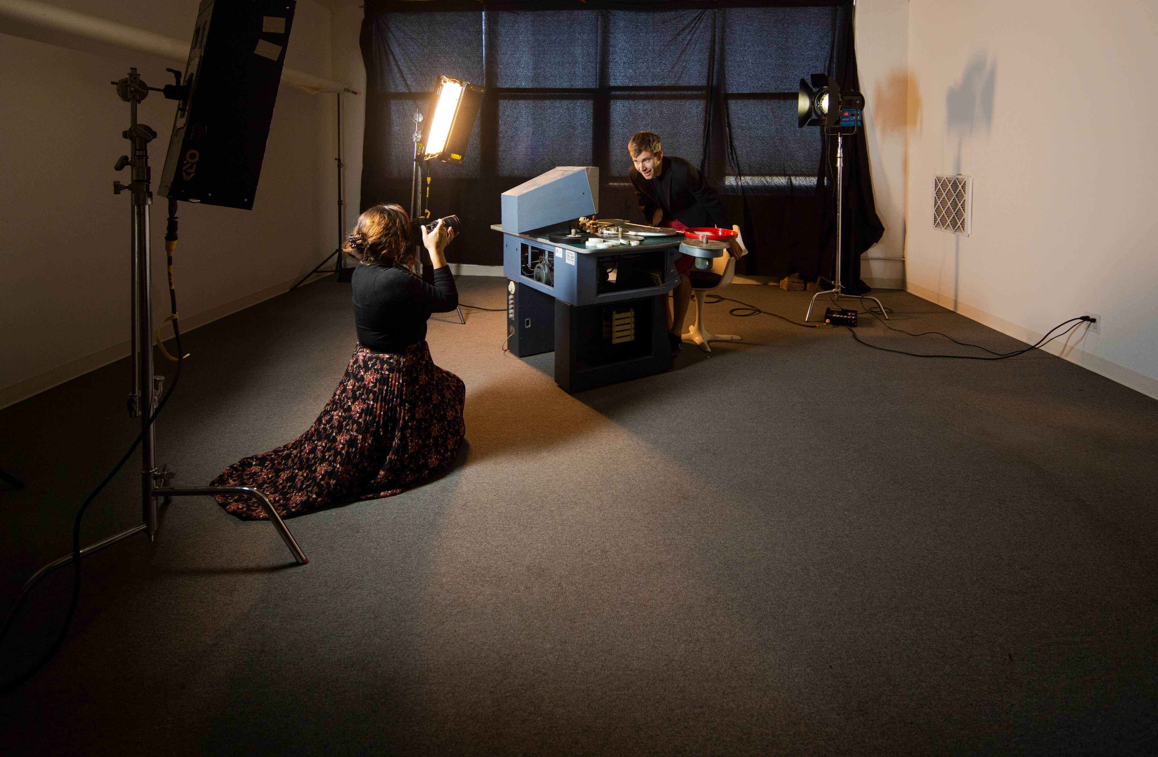 A woman sits on the floor behind a camera while a man operates a machine in a well-lit studio space.