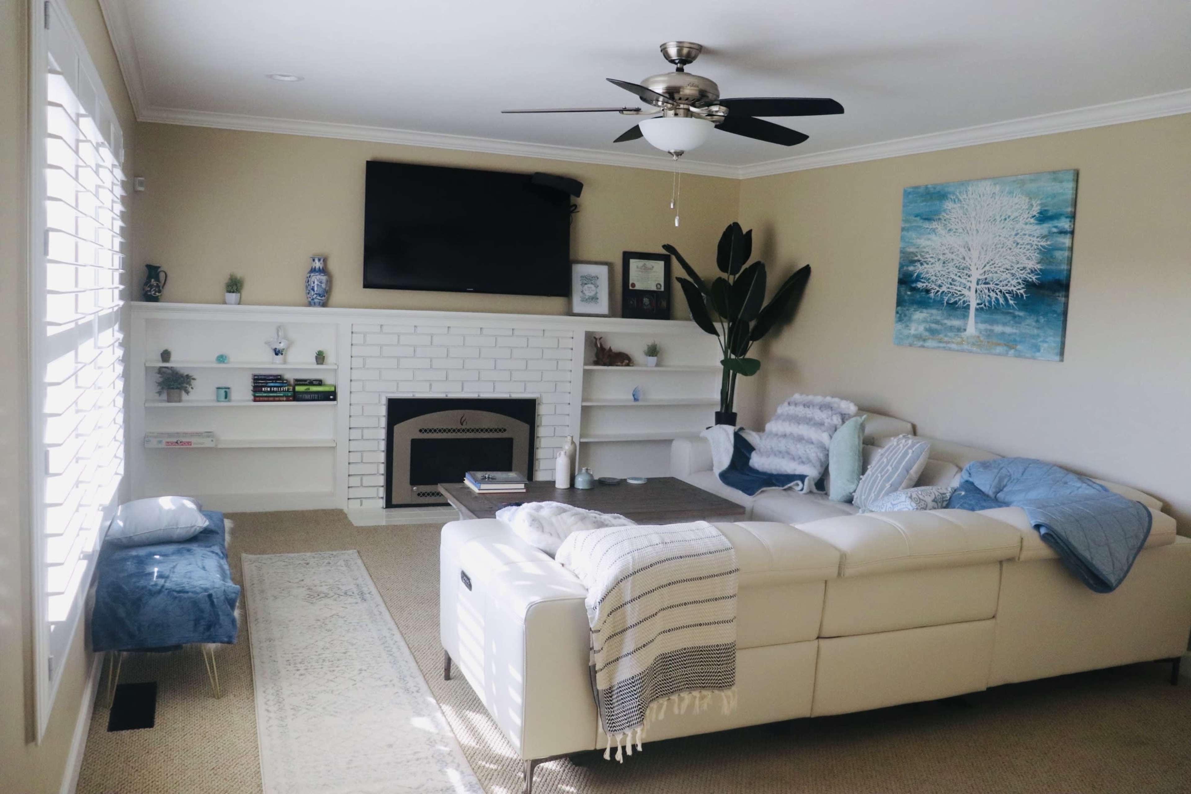 A bright living room features a white sectional sofa, a ceiling fan, a flat-screen TV mounted above a white brick fireplace, and a blue and white artwork on the wall.