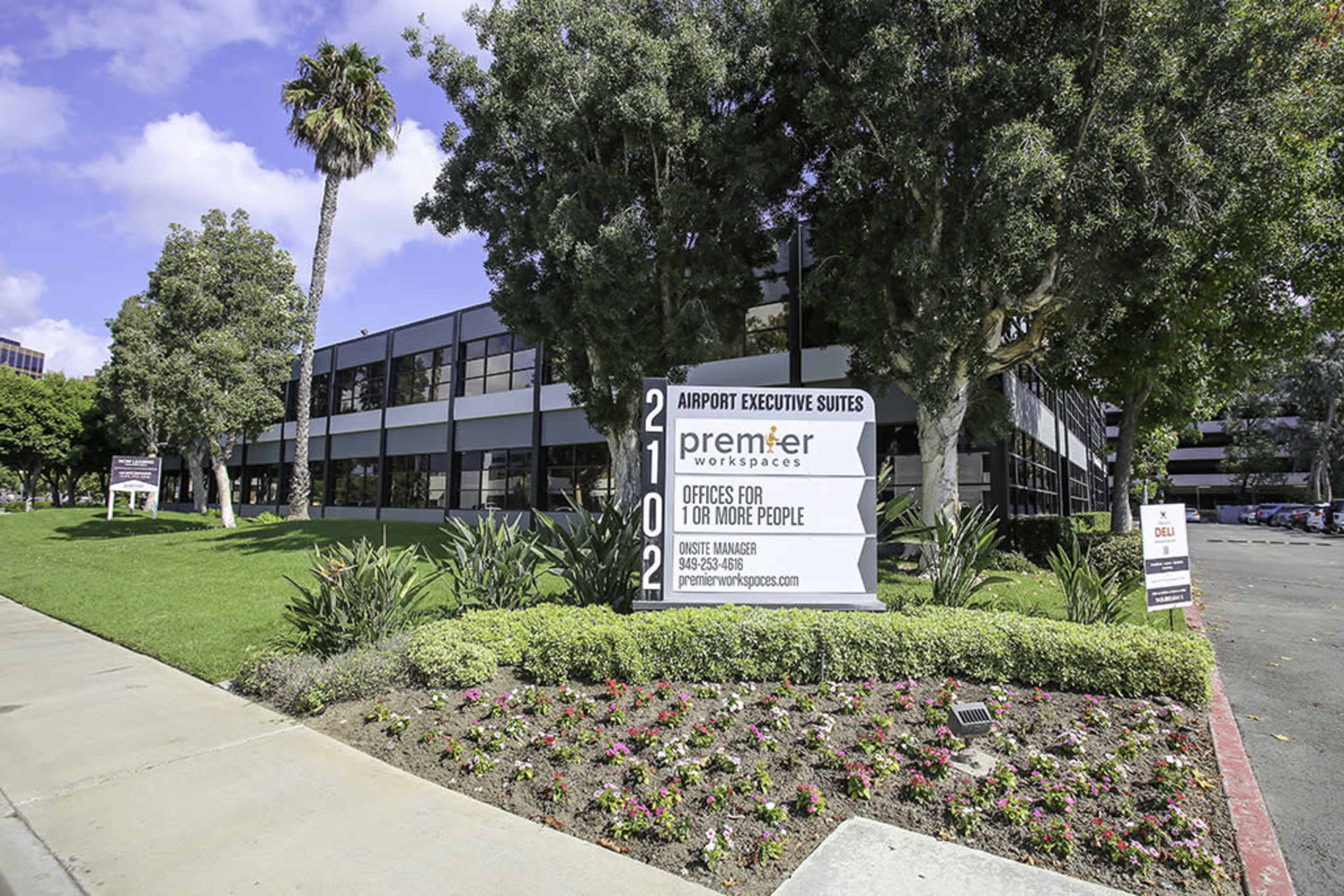 The image shows a multi-story commercial building named "Airport Executive Suites," with a sign indicating available office spaces for one or more people, surrounded by landscaped greenery and flowers.