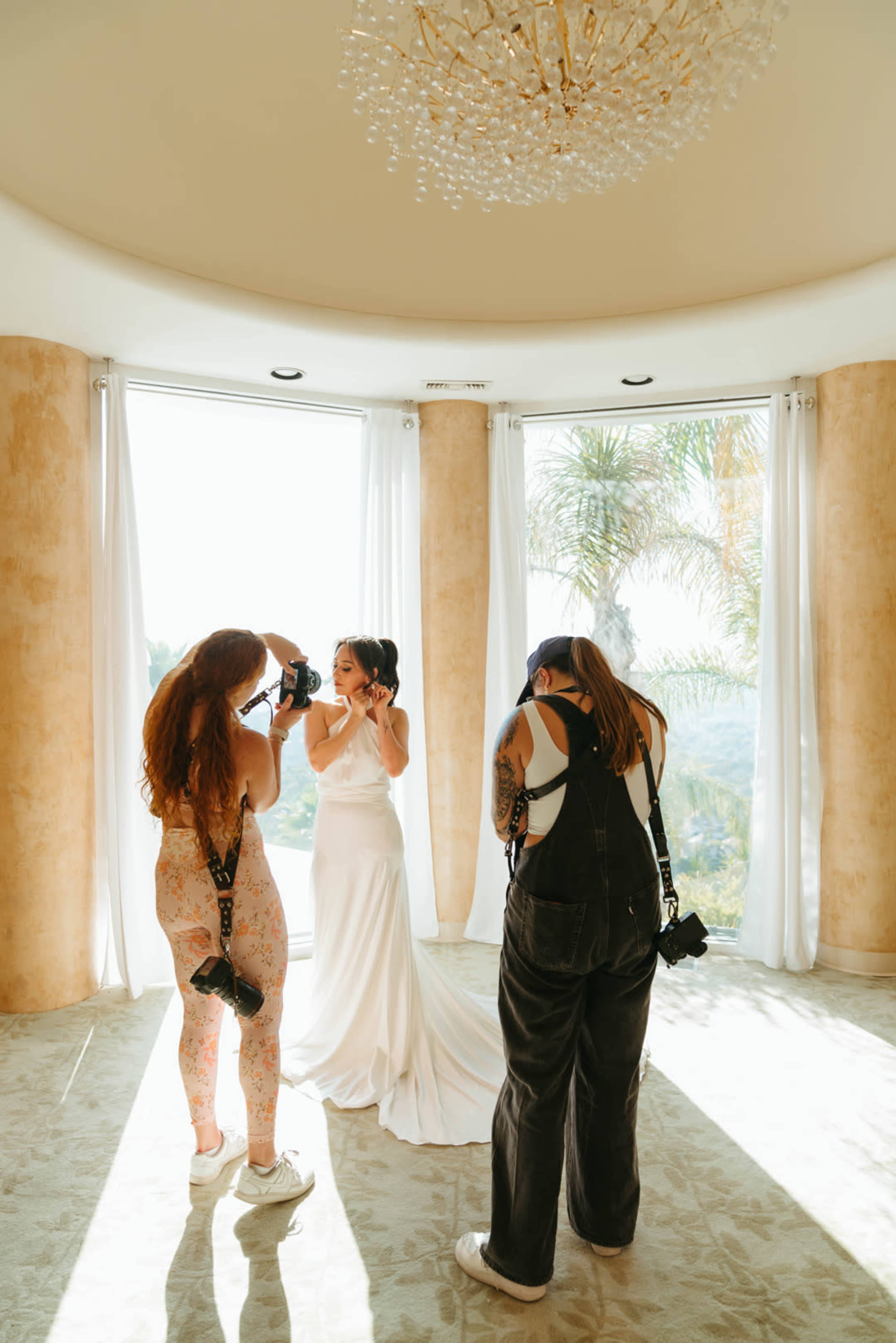 A bride is getting her makeup done while two women photographers capture the moment in a brightly lit room with large windows.