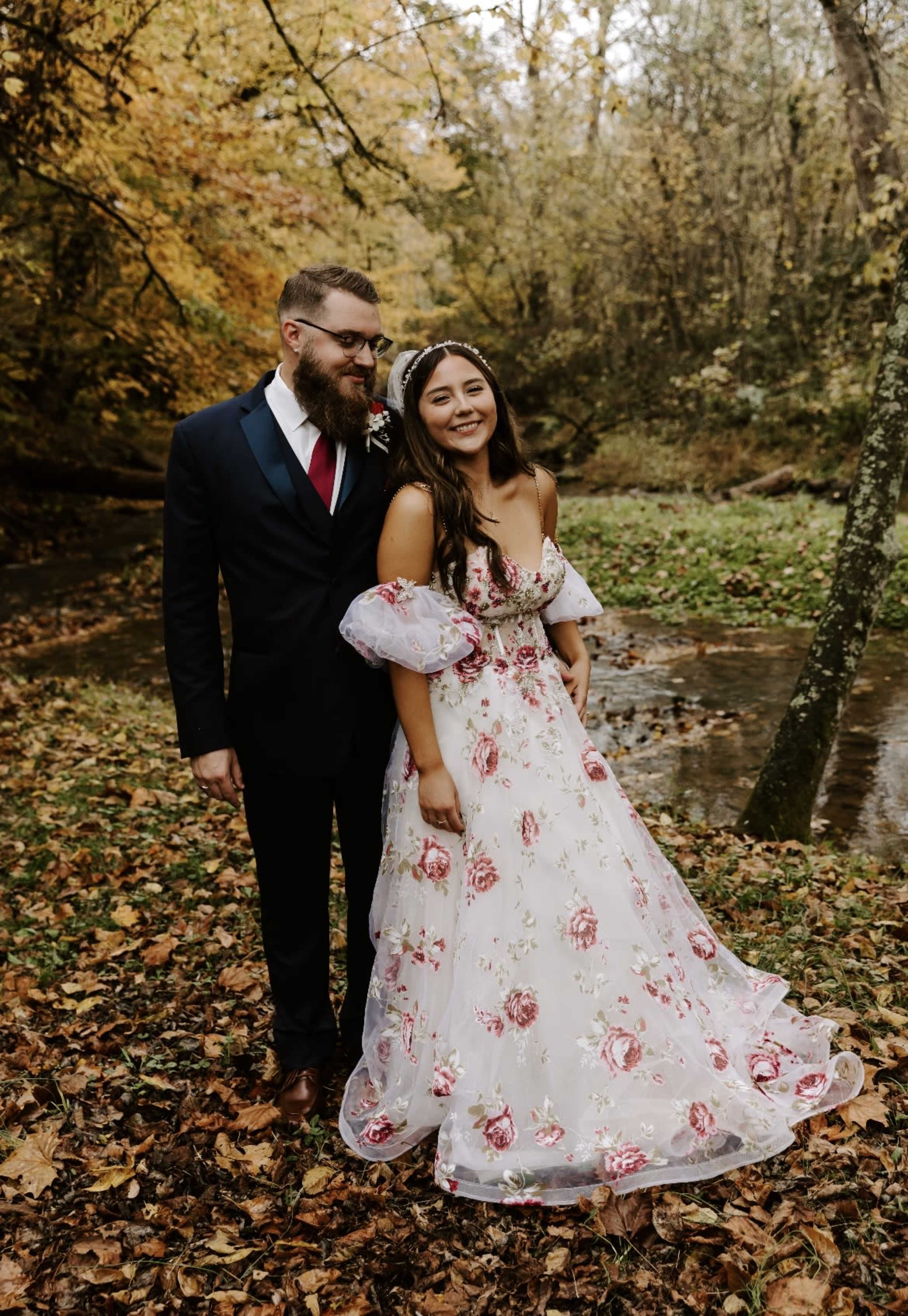 A couple stands together in a forest clearing adorned with autumn leaves, with the woman in a floral wedding dress and the man in a dark suit.