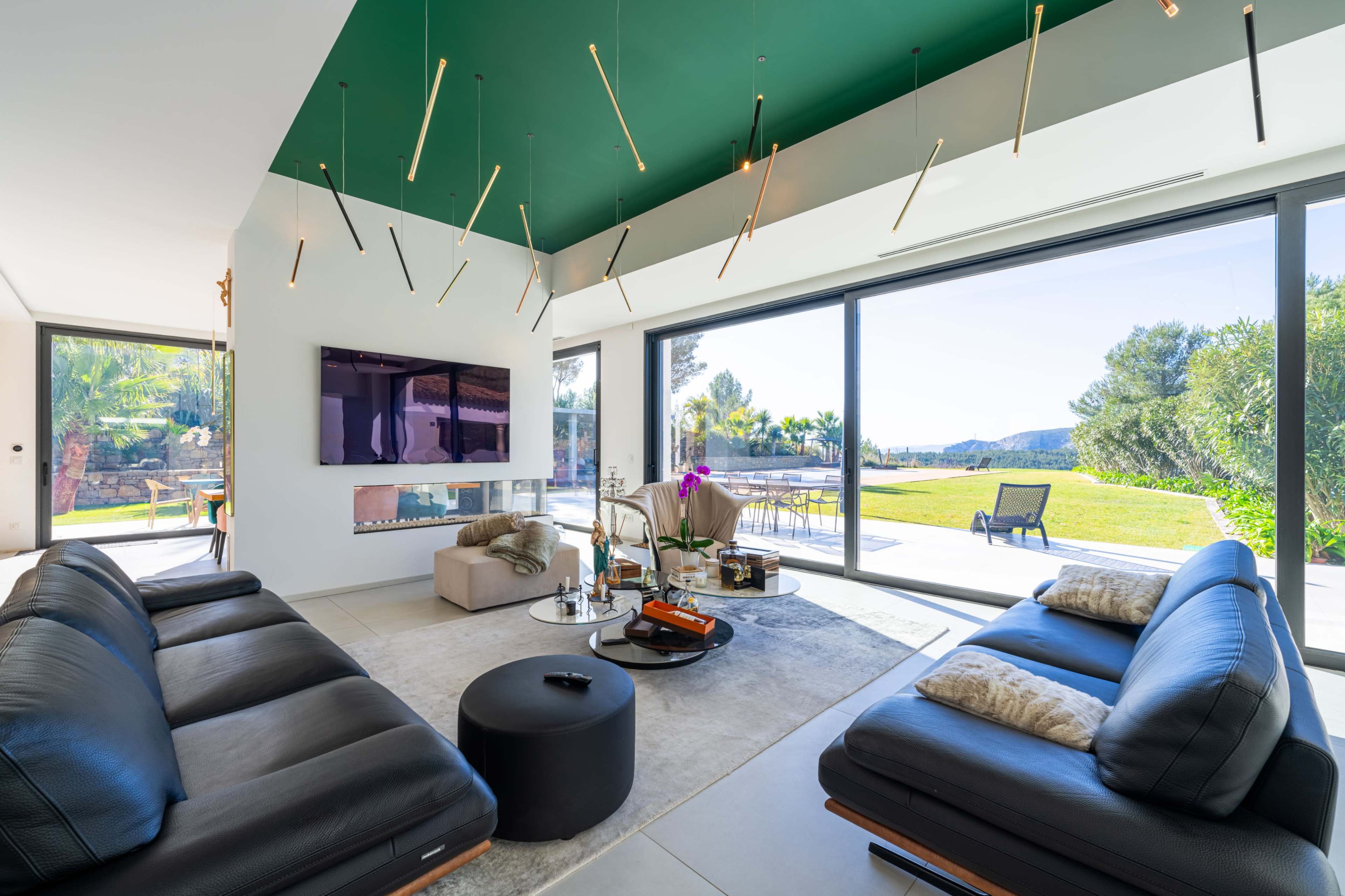 The image shows a modern living room with black leather sofas, a television on the wall, and large glass doors opening to a green outdoor space with trees.