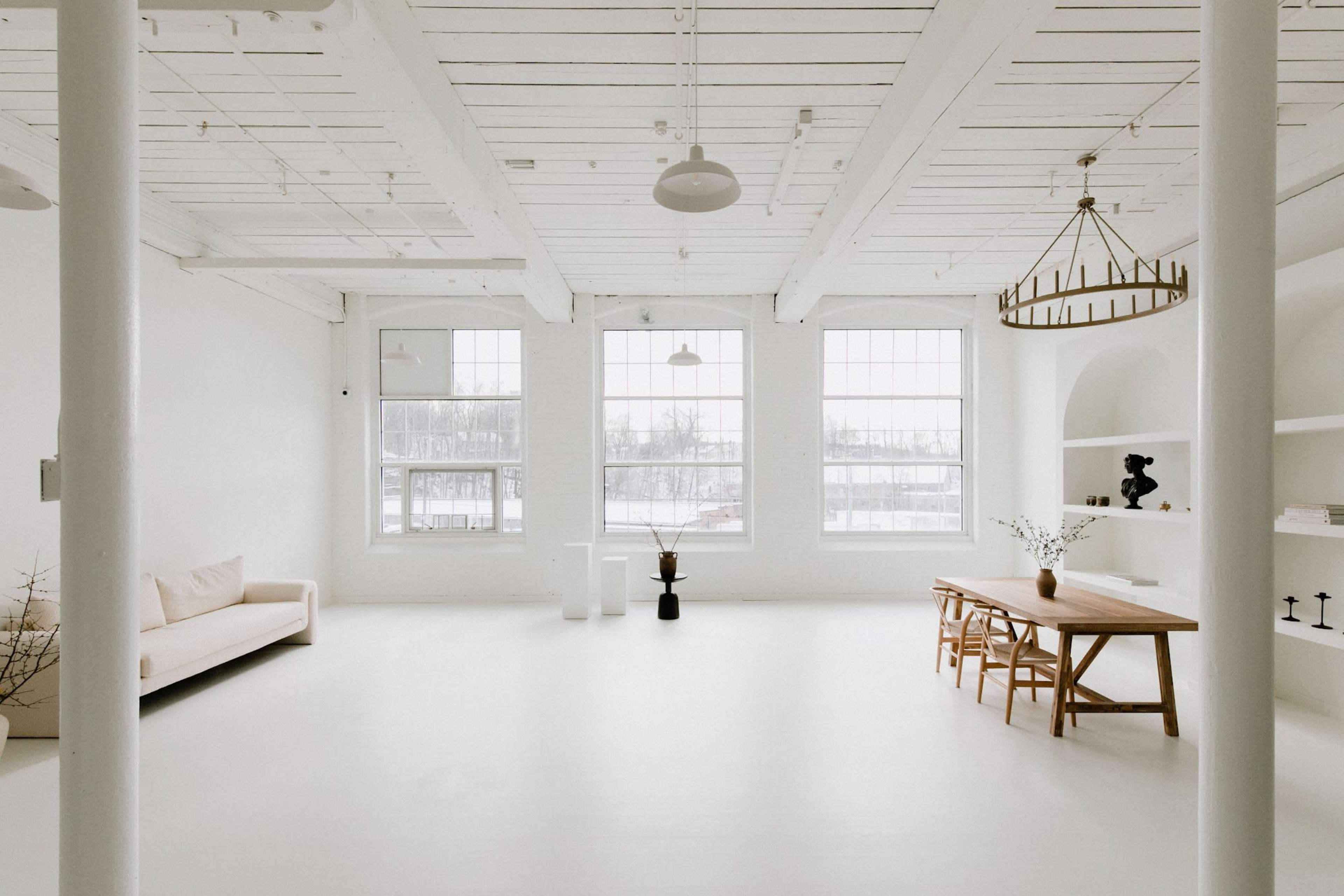 A bright, minimalist interior space featuring a sofa, a wooden dining table, and large windows surrounded by white walls and ceiling beams.