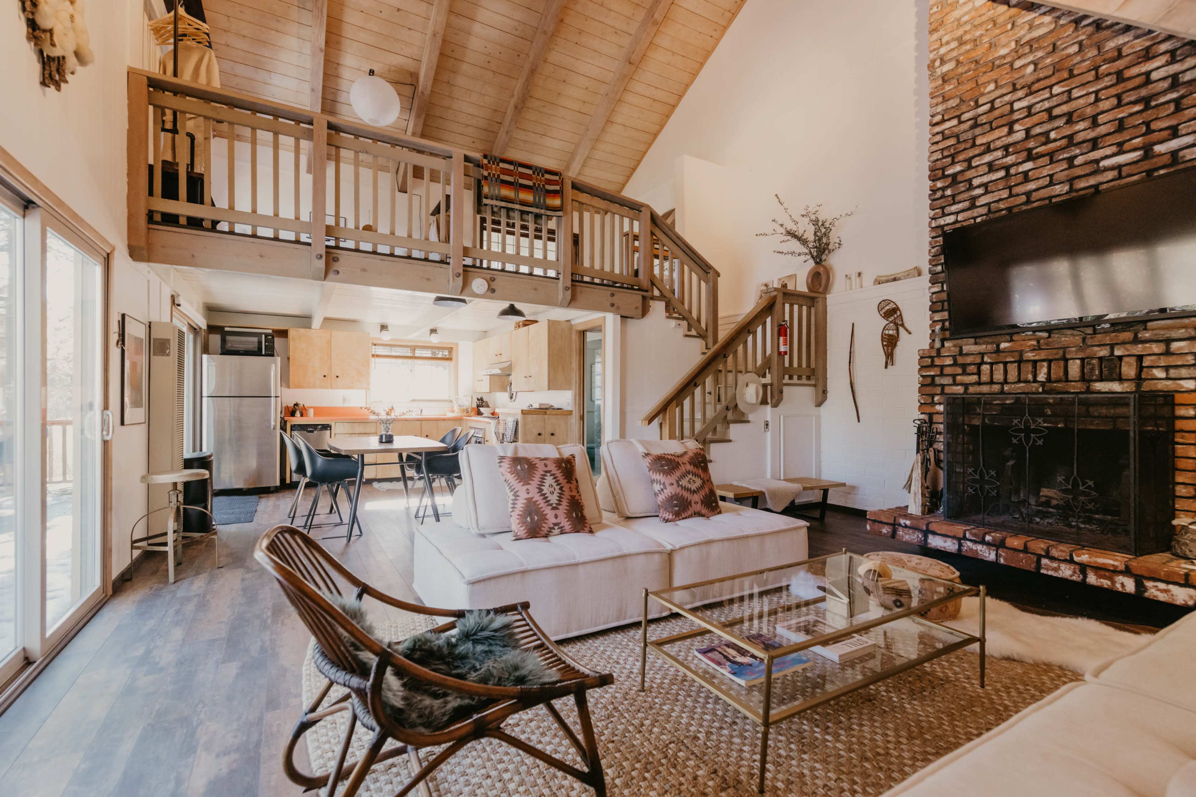 A cozy living room features a white sectional sofa, a glass coffee table, and a large brick fireplace, with a staircase leading to a lofted area above.