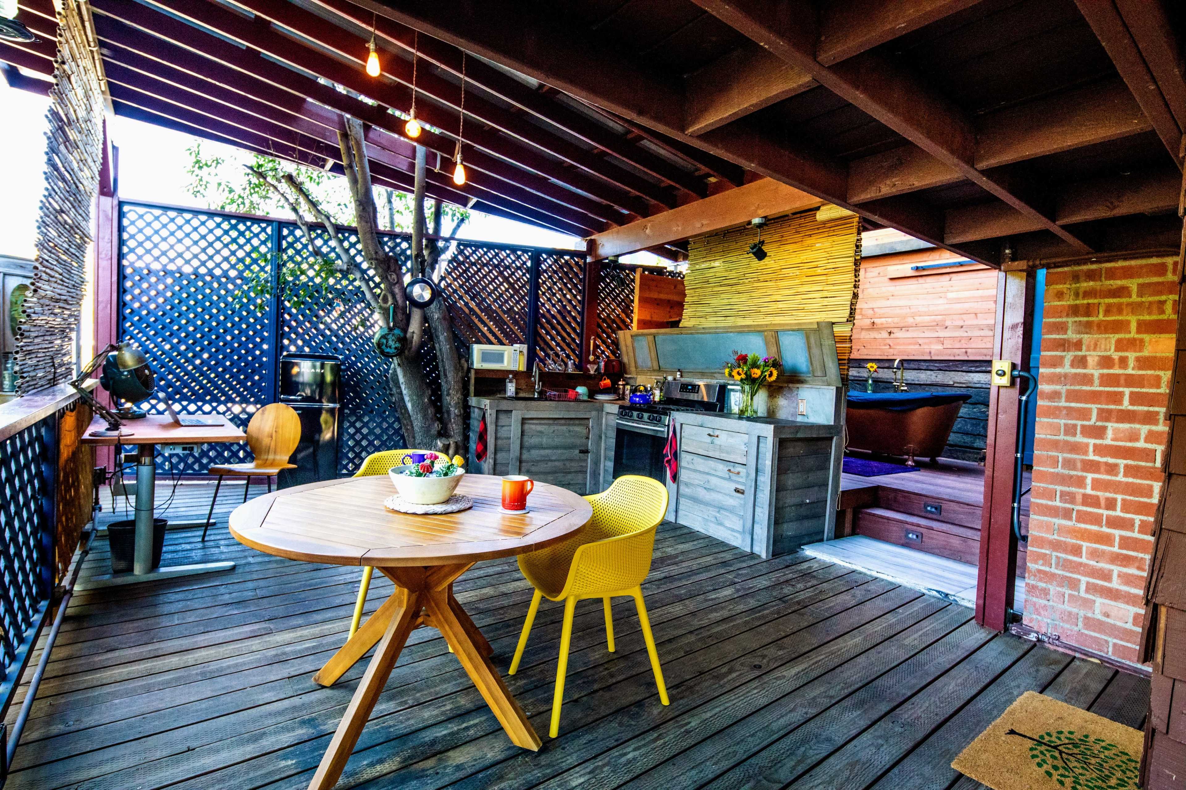The image shows an outdoor dining area with a round wooden table, yellow chairs, and a kitchen setup featuring a grill and countertop under a wooden structure.