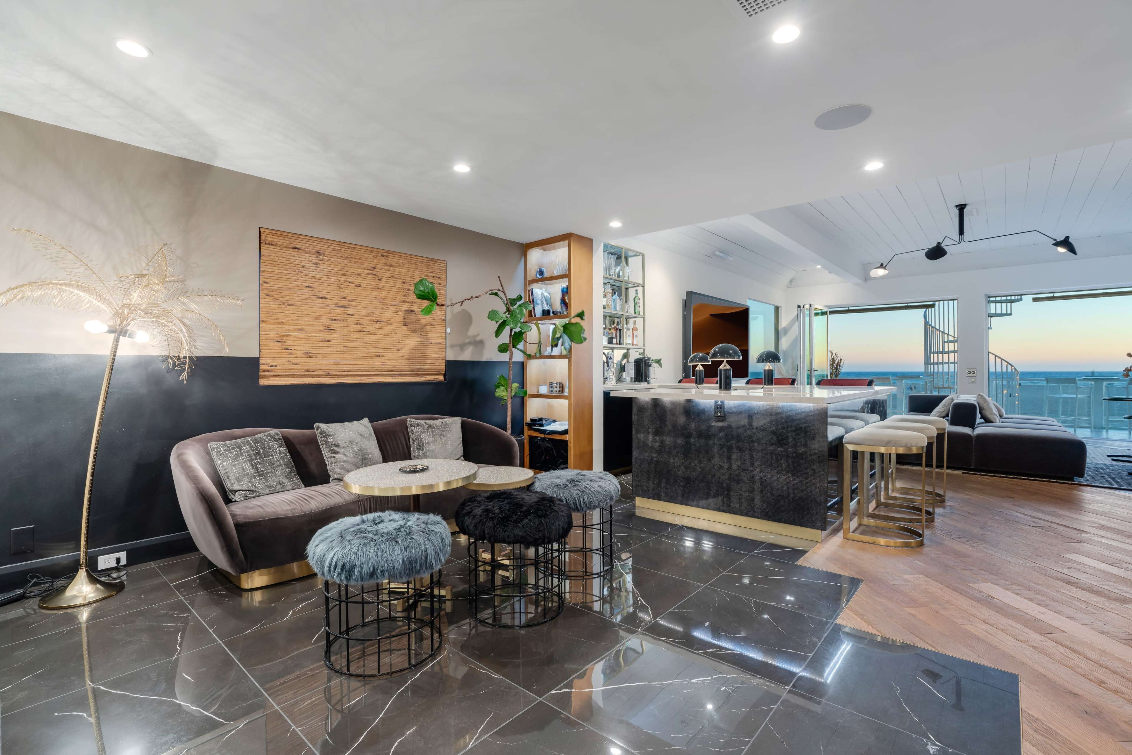 A modern living space featuring a plush sectional sofa, decorative tables, and a kitchen area with bar seating, all set against large windows displaying a sunset view.