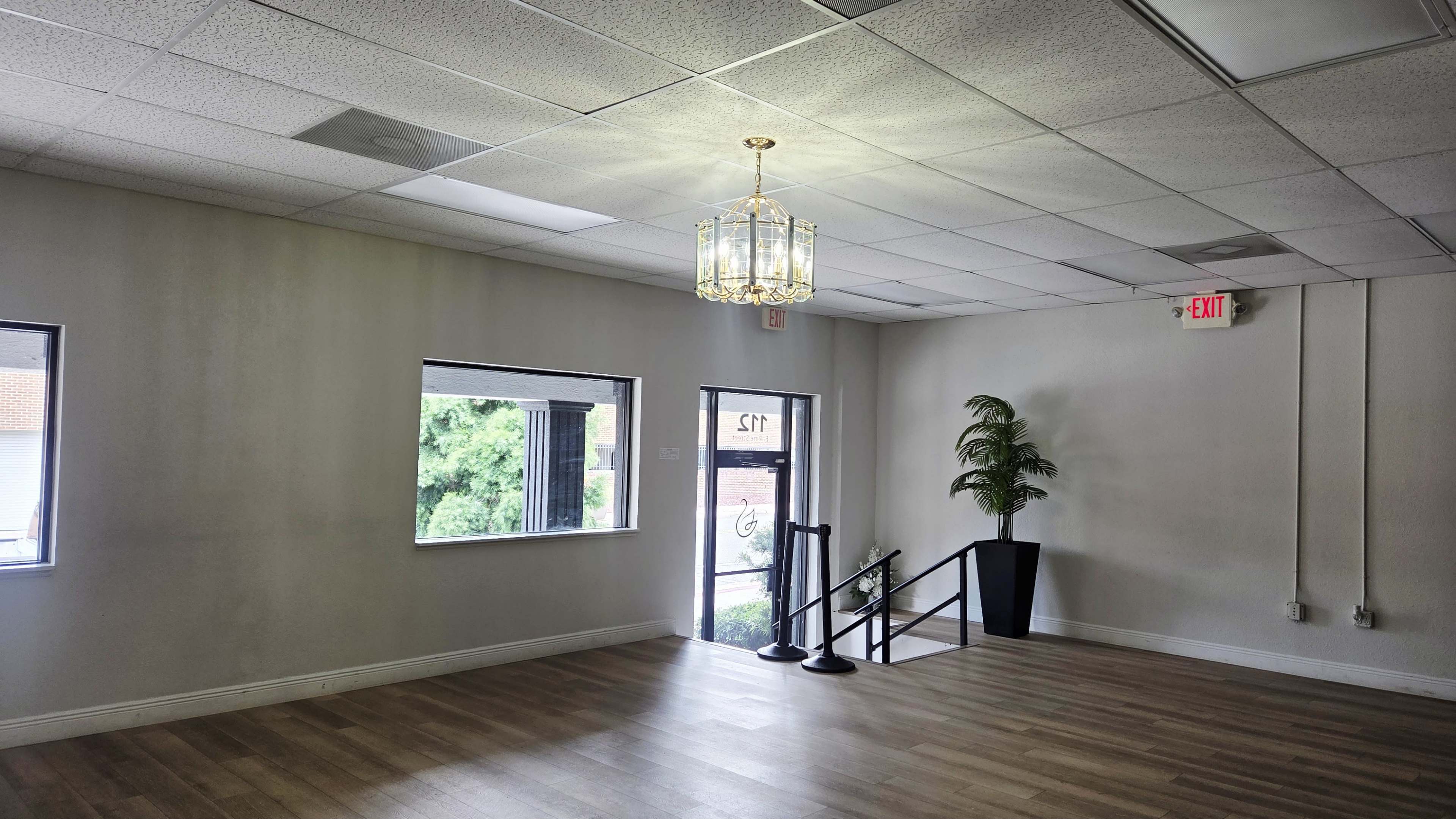 The image shows a vacant room with wooden flooring, a chandelier, and large windows, featuring a staircase leading to the entrance.