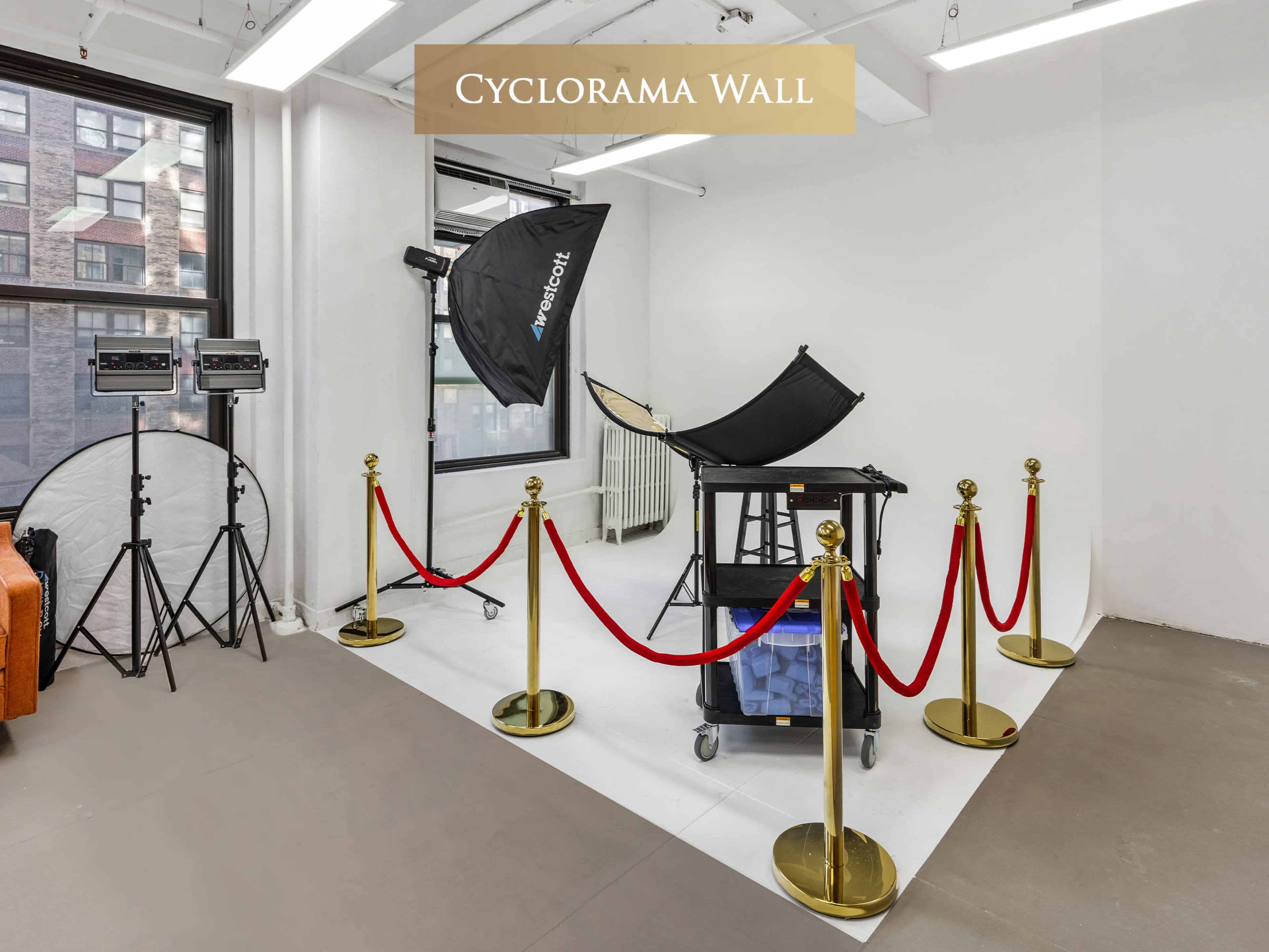 The image shows a photography studio featuring a cyclorama wall, equipped with a black backdrop, lighting equipment, and a cart with various items, all surrounded by red velvet ropes and gold stanchions.