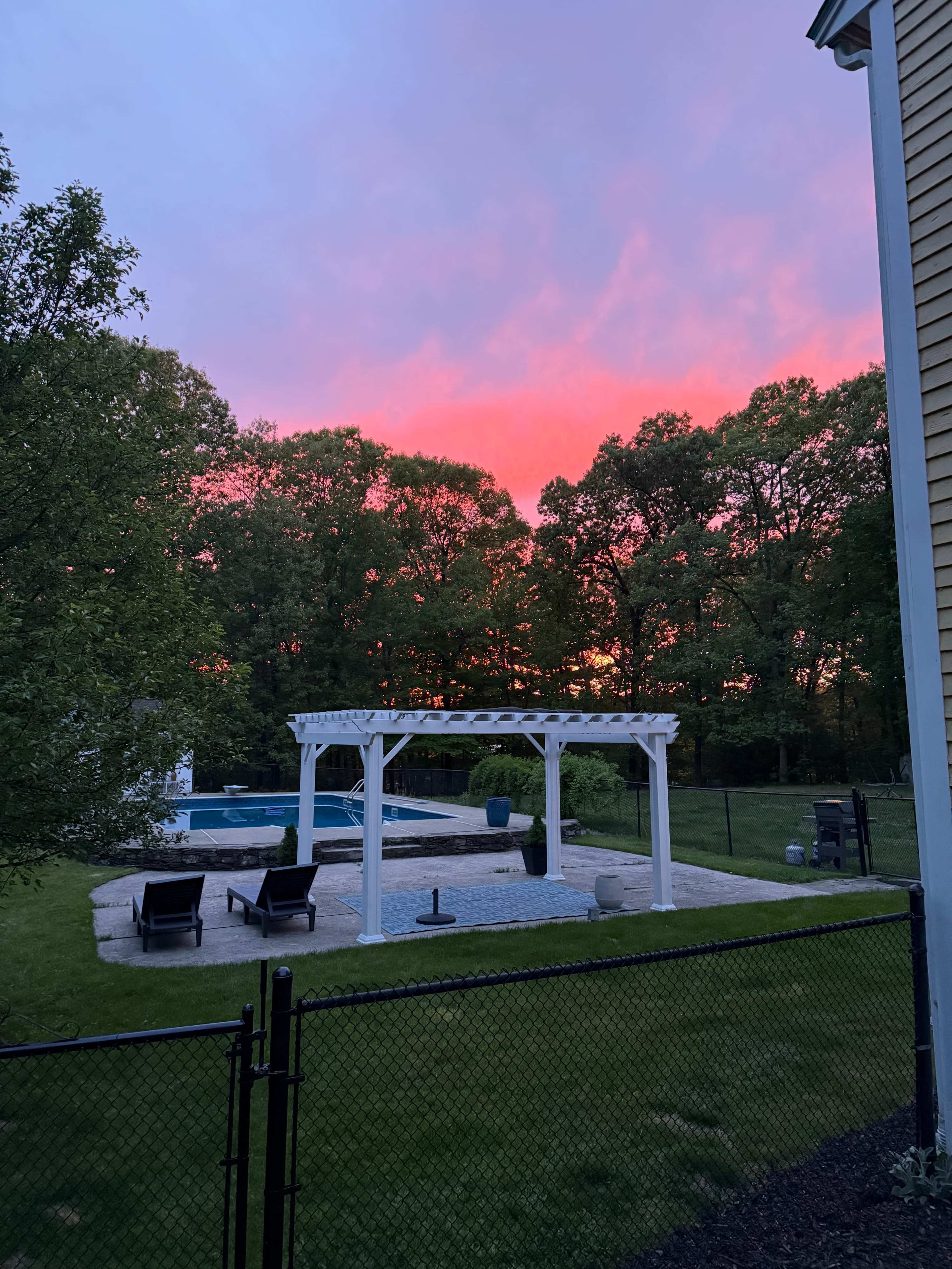 A backyard features a pool area with lounge chairs and a white pergola, set against a vibrant sunset sky.