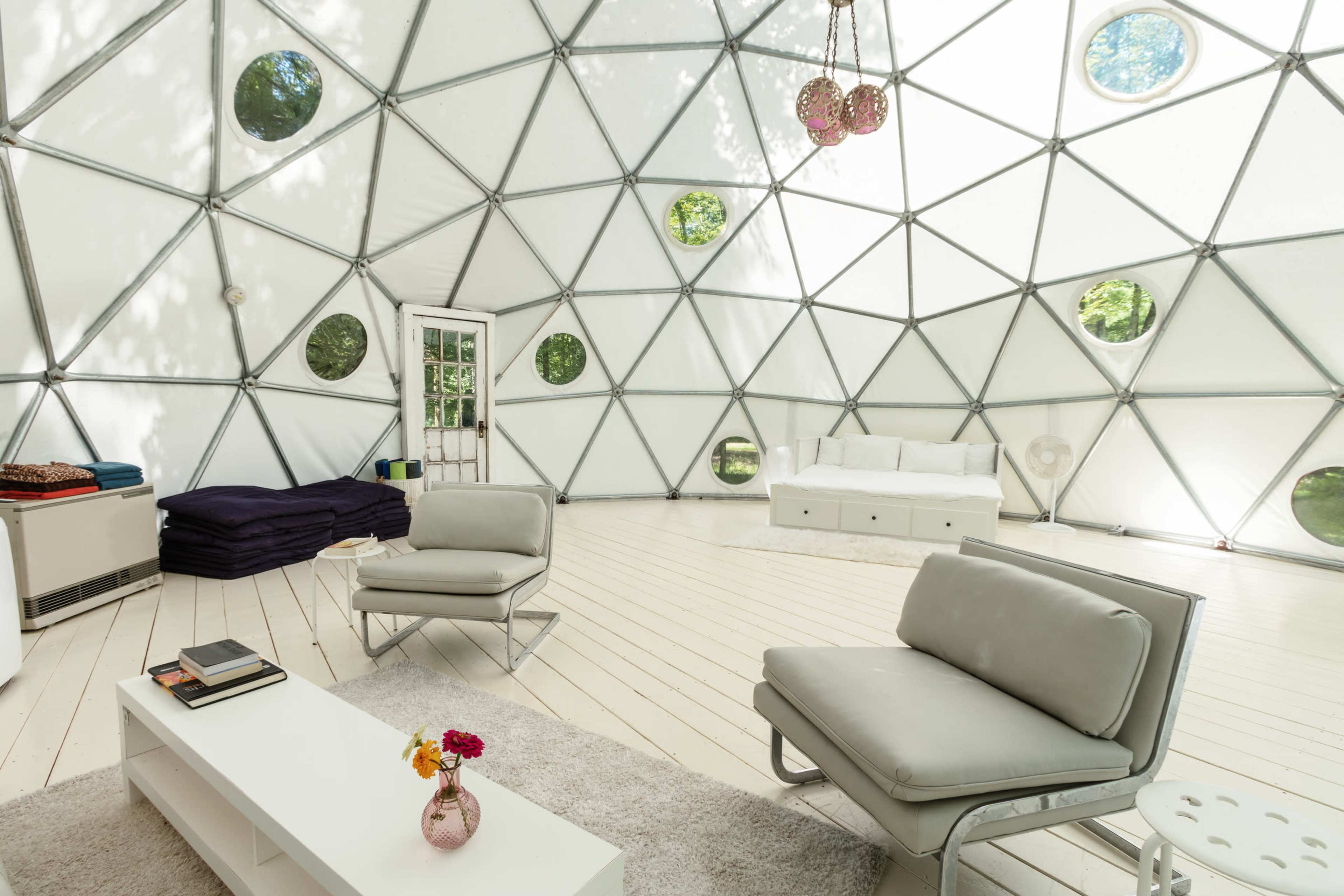 The interior of a geodesic dome features modern furniture, a white rug, and large windows allowing natural light to fill the space.