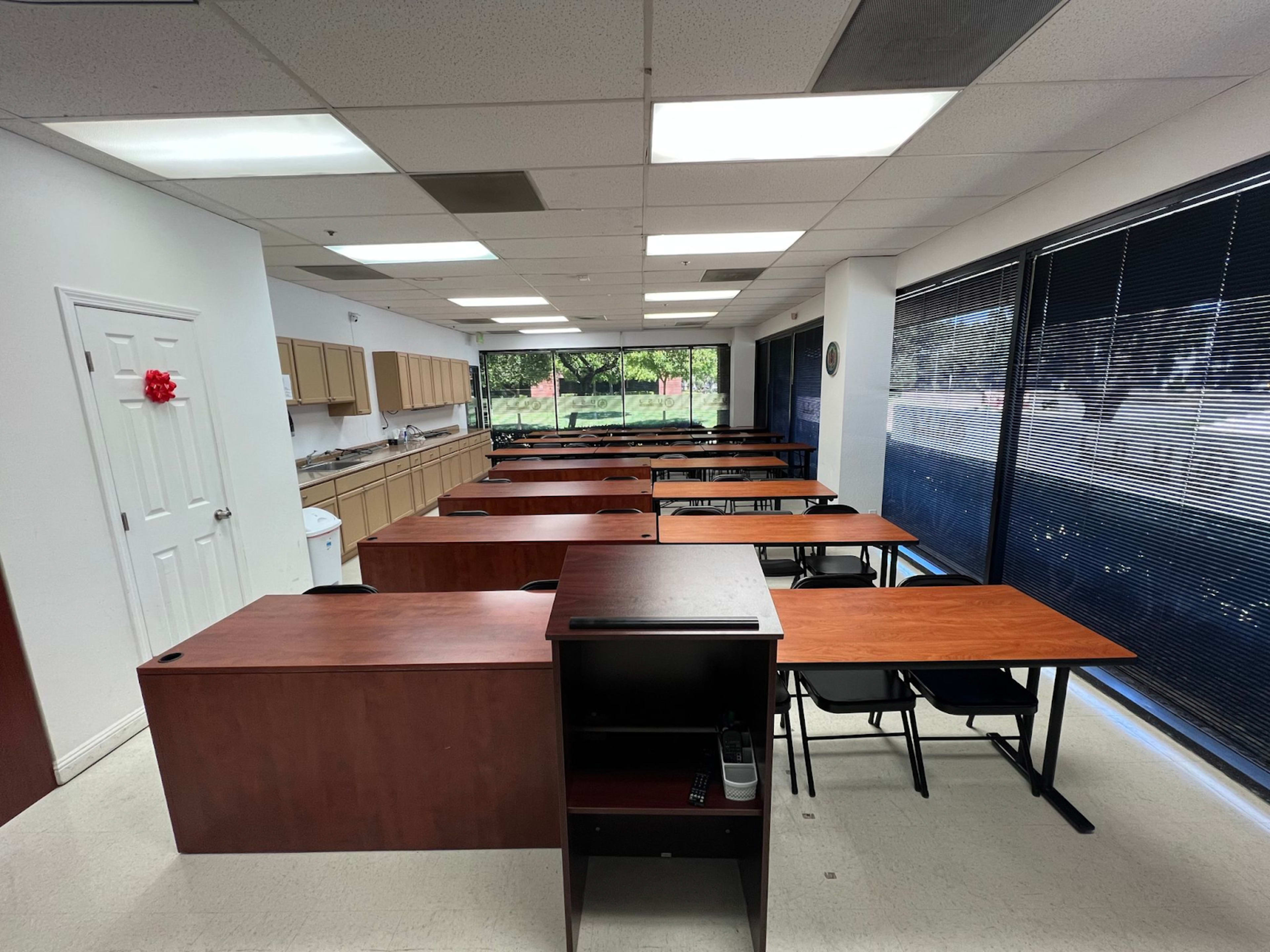 The image shows a classroom setup with rows of wooden desks and a kitchen area in the background, illuminated by natural light from large windows.