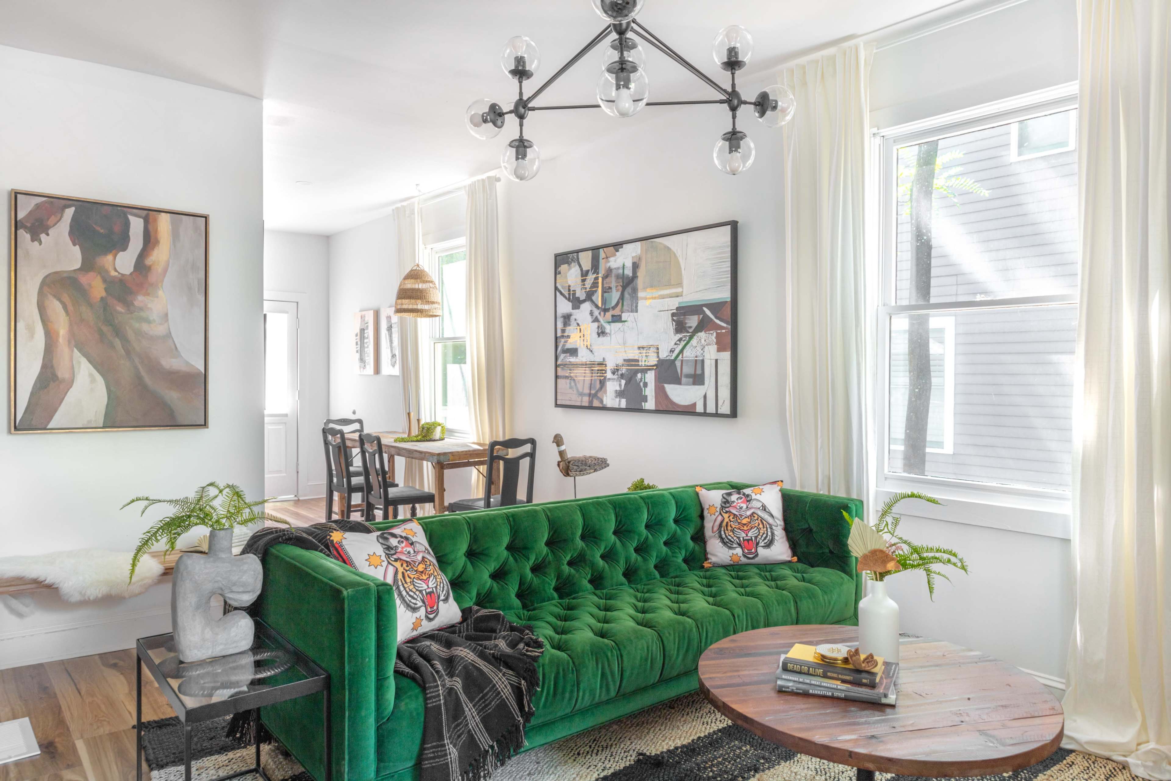 A modern living area featuring a green velvet sofa with decorative pillows, a round wooden coffee table, and large windows allowing natural light to fill the space.