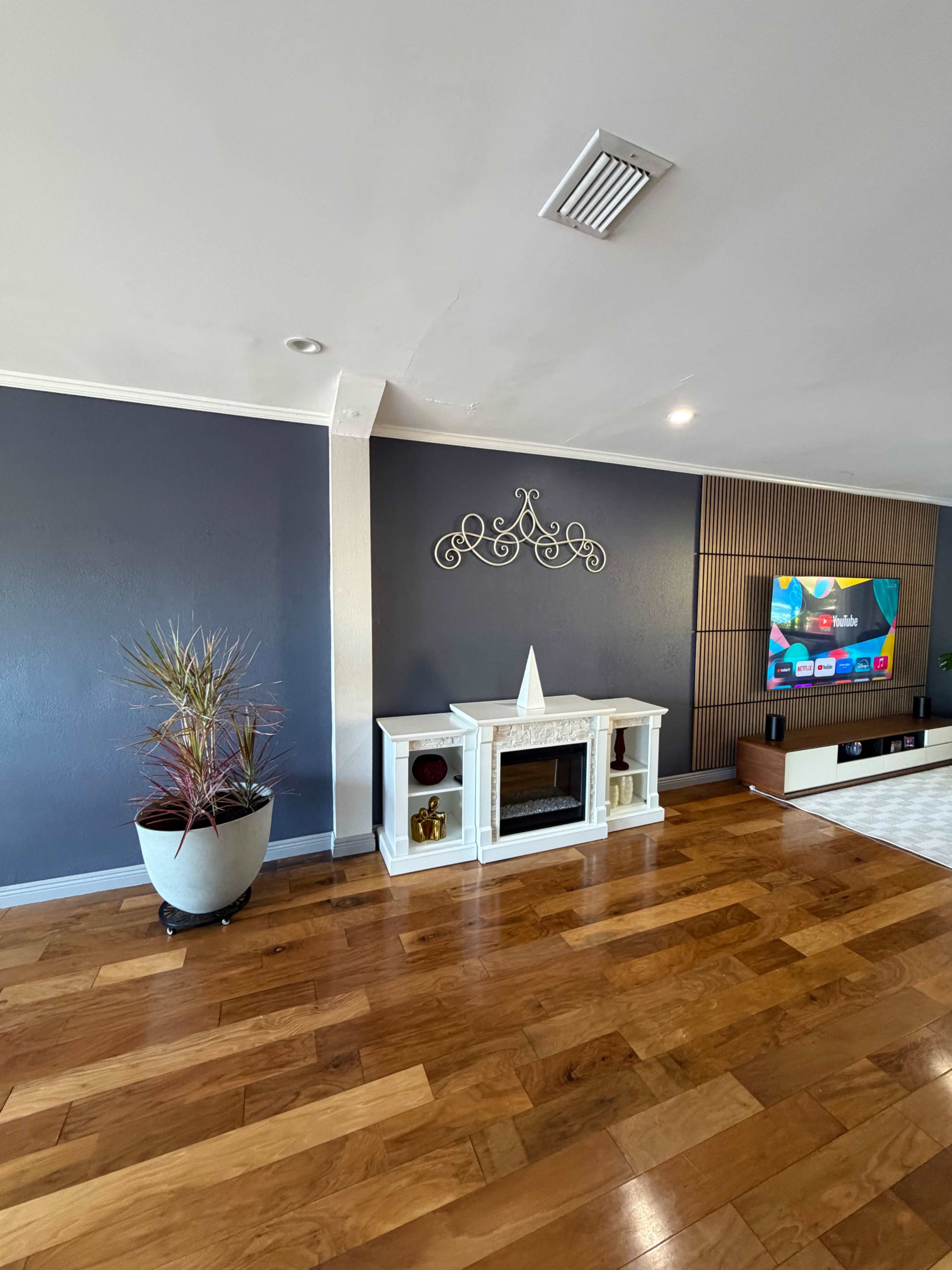 The image shows a modern living room with a fireplace, a large television mounted on the wall, wooden flooring, and a potted plant in the corner.