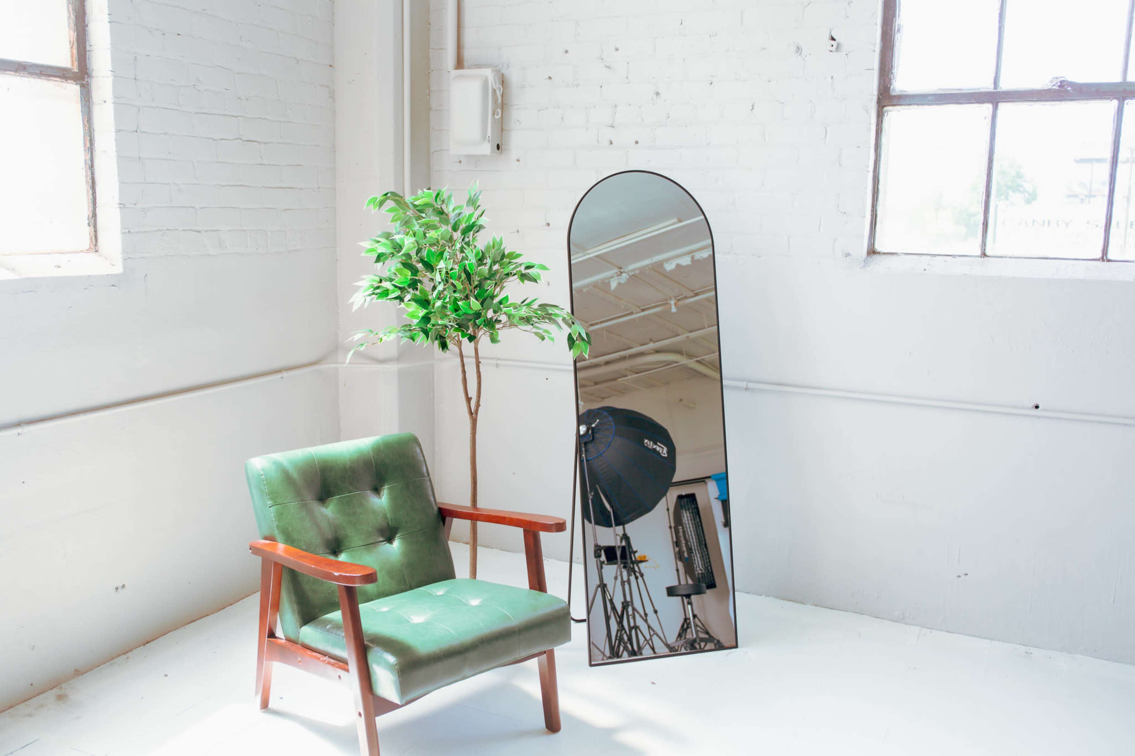 The scene features a green upholstered chair next to a tall mirror and a potted tree in a bright, minimalist room with windows.