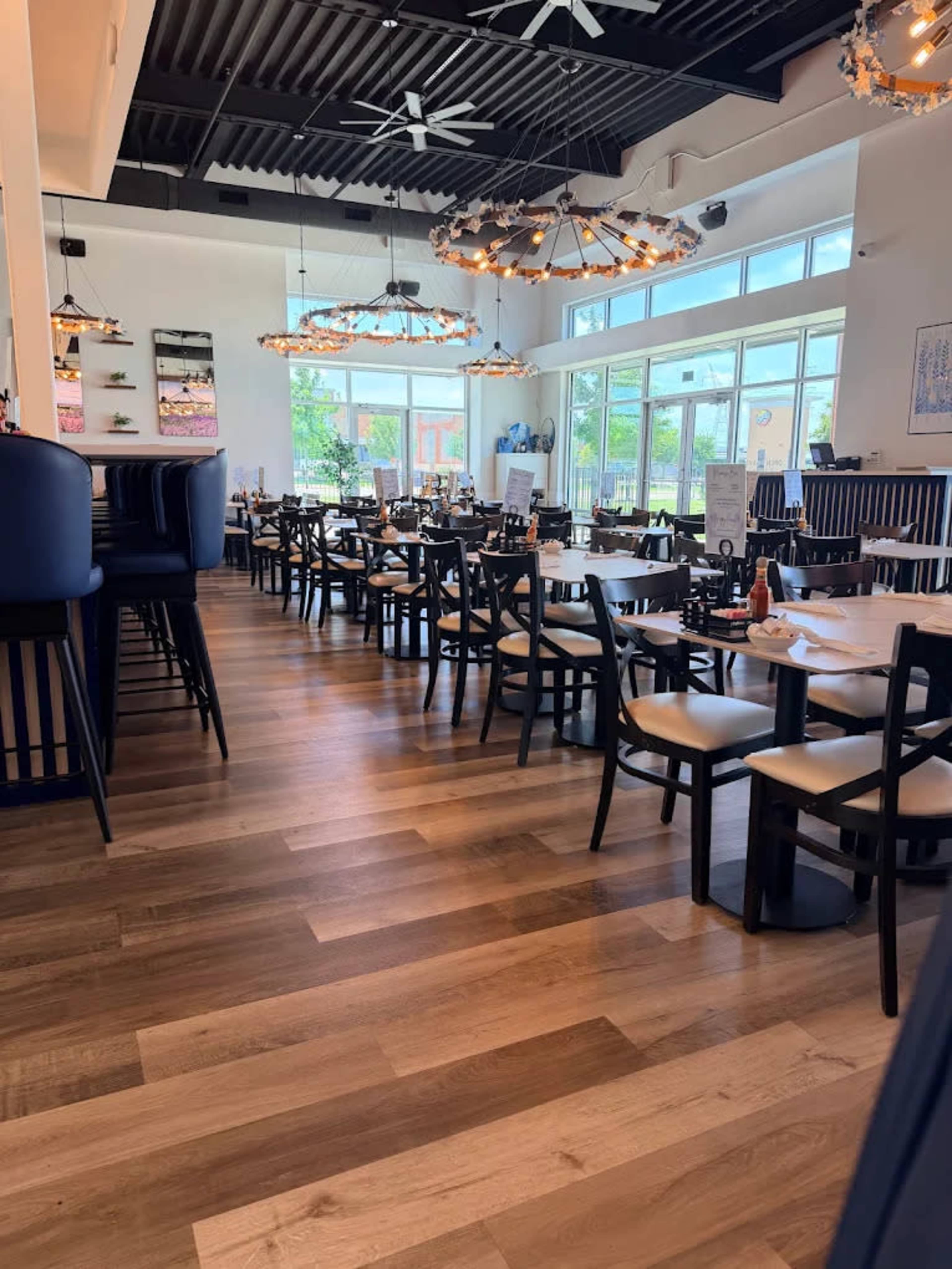 The image shows a modern restaurant interior with wooden flooring, tables set for dining, and large windows allowing natural light to enter.