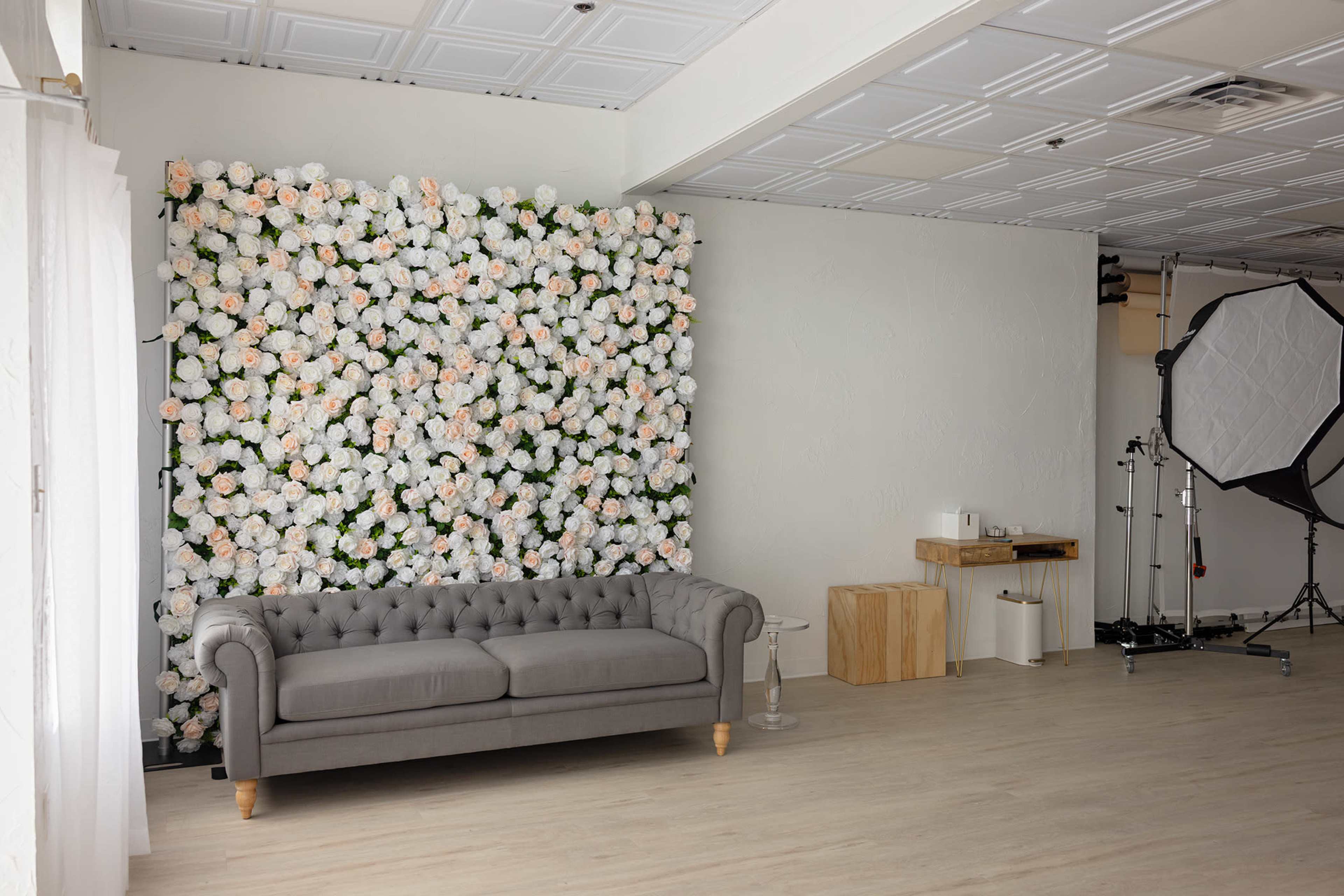 The image shows a photography studio featuring a tufted gray couch in front of a large wall adorned with artificial white and peach flowers.