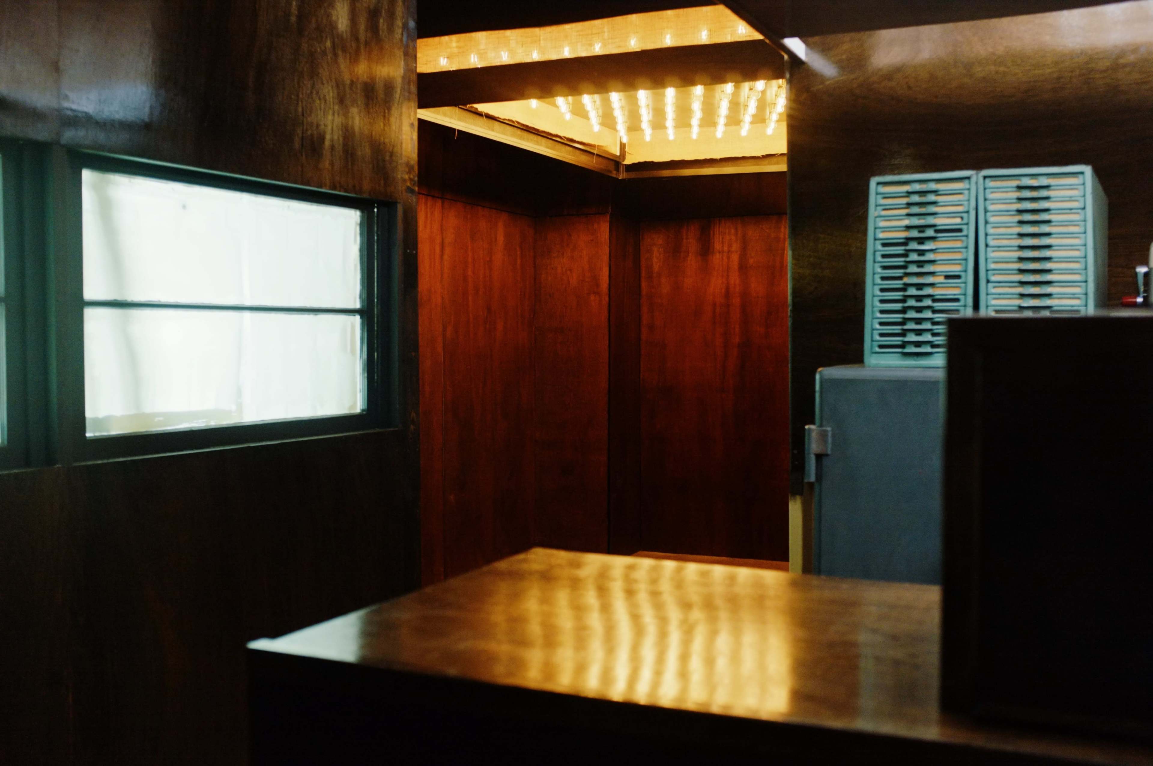 A dimly lit interior space with wooden walls, a glass window, and a doorway leading to a well-lit area.