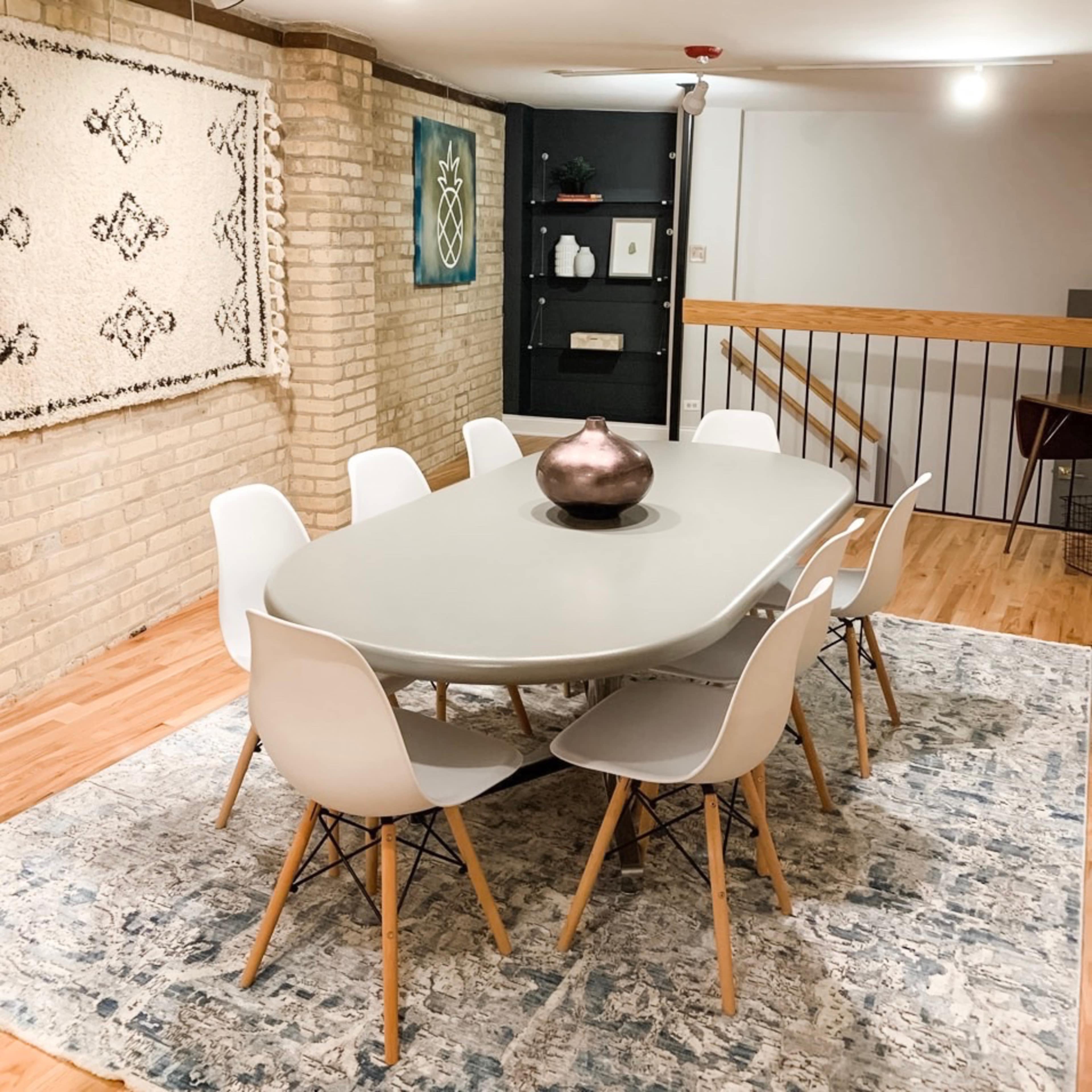 A modern dining area features a large oval table surrounded by six white chairs on a patterned rug, with a brick wall and decorative elements in the background.
