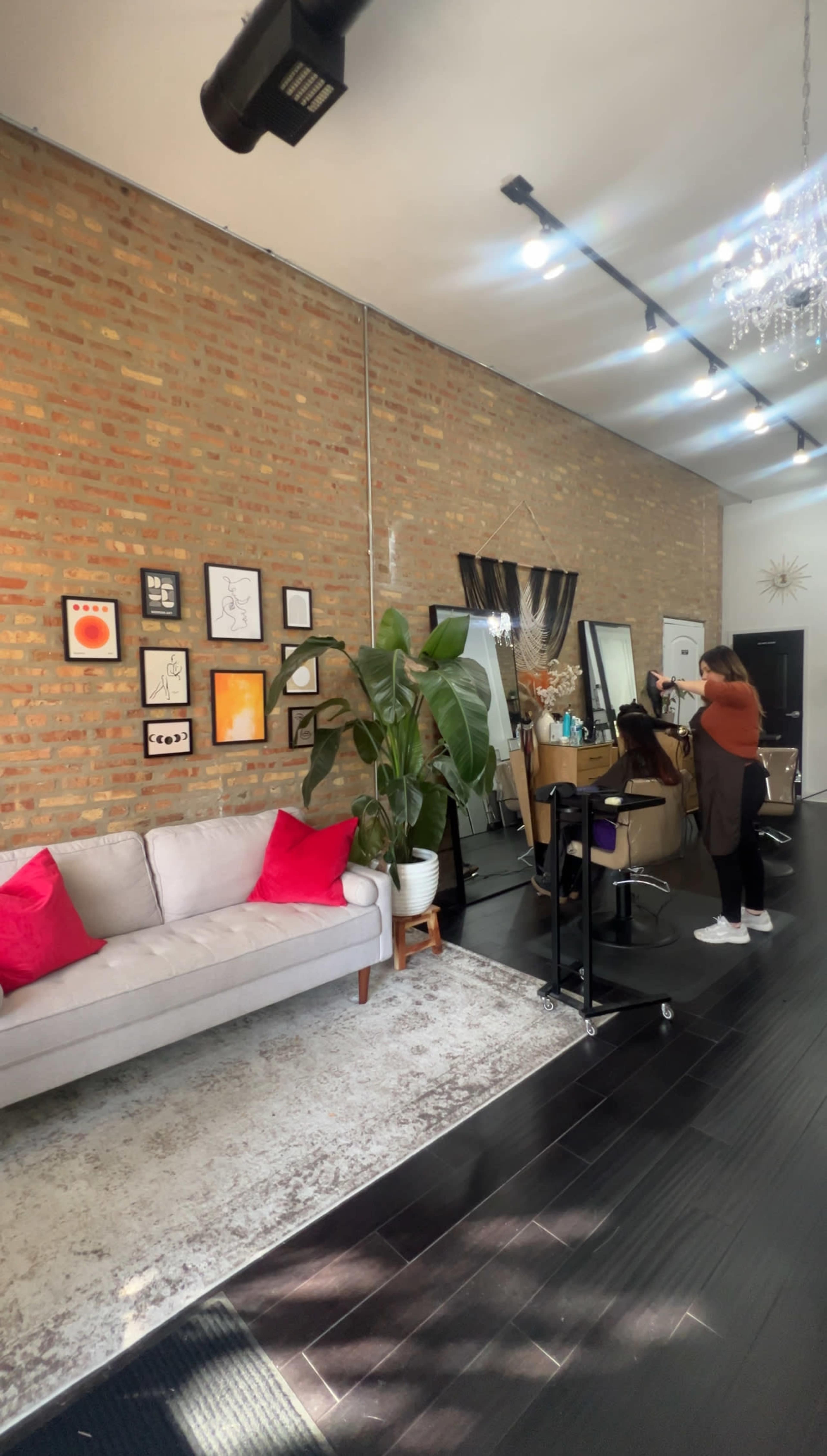 The image shows a modern hair salon with a brick wall, a gray couch with red cushions, and a stylist working at a salon station.