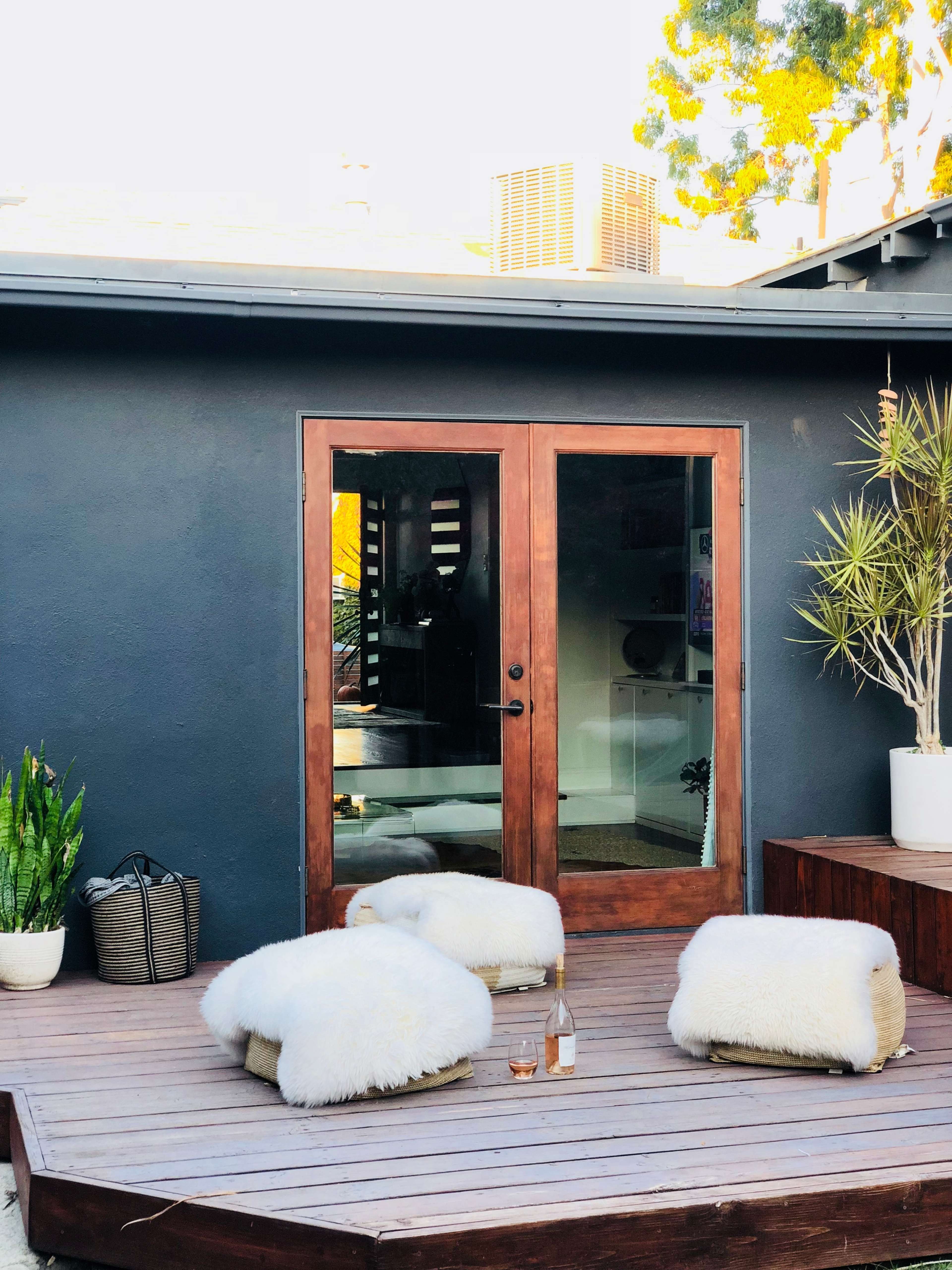 A wooden deck with three white poufs, a bottle of rosé wine, and potted plants, in front of a glass door leading into a house with dark walls.