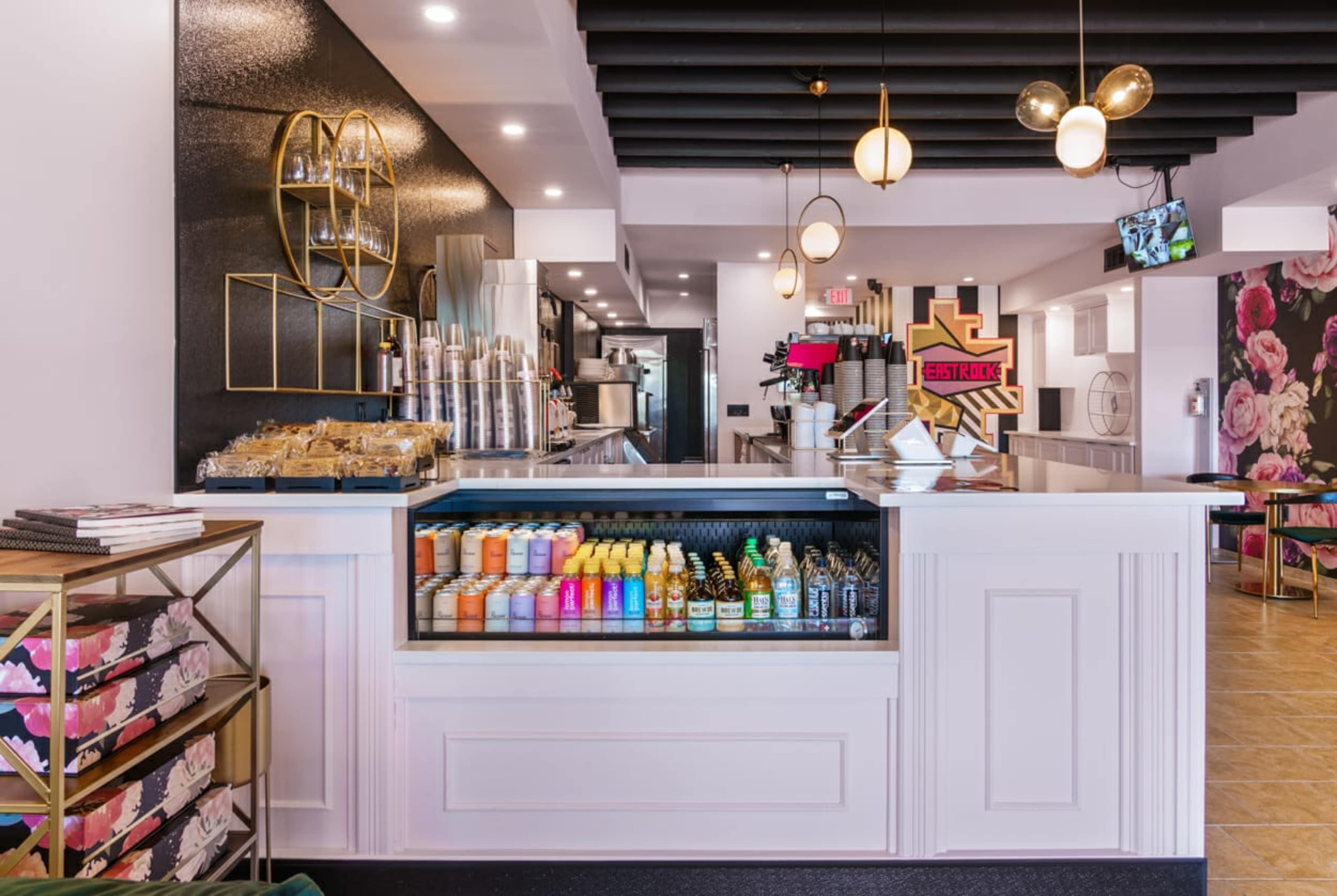 A modern café interior features a sleek counter displaying a variety of colorful drinks and pastries, with stylish lighting and floral decor in the background.
