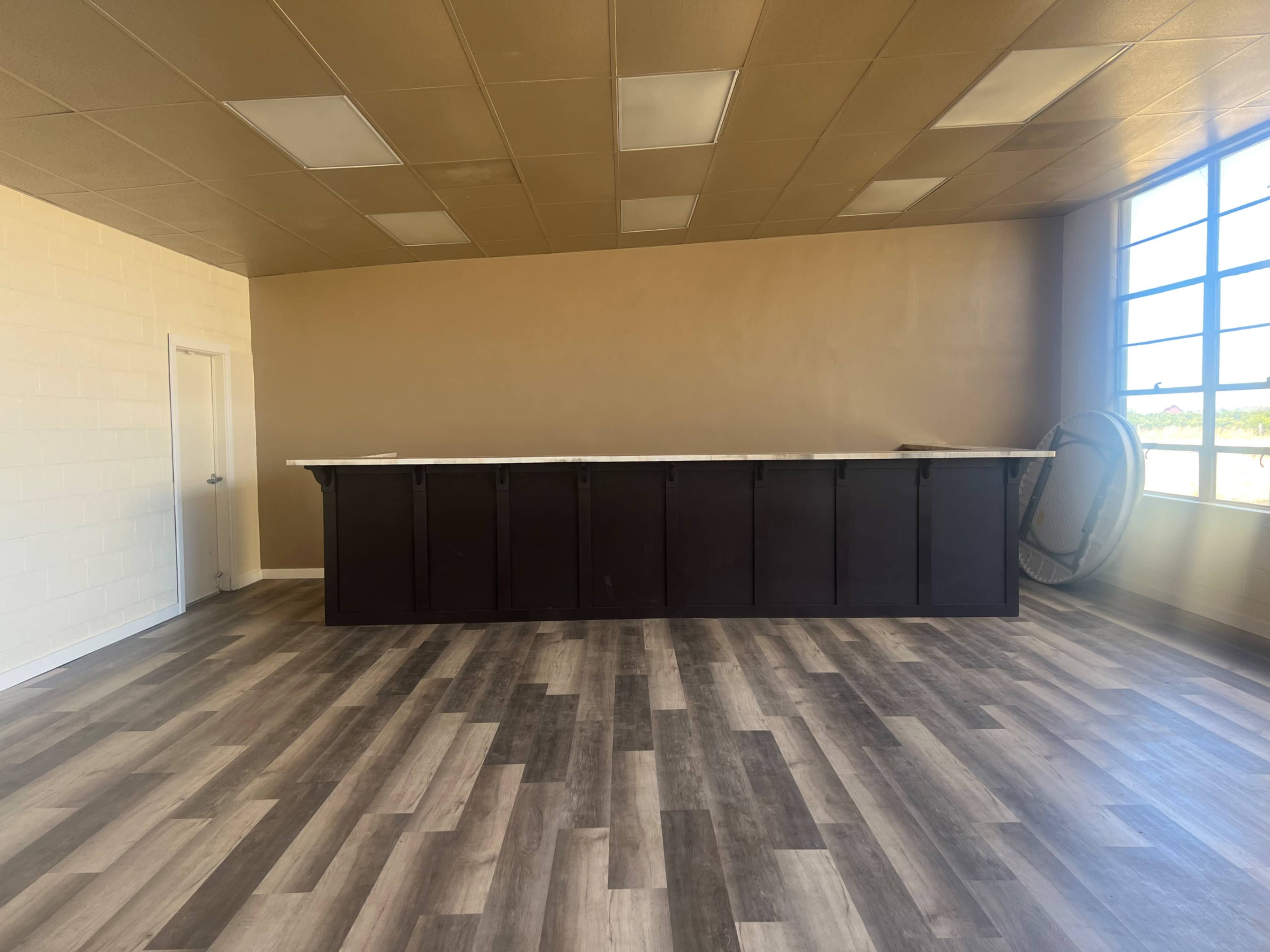 An empty room with a wooden floor, a long dark counter against the back wall, and large windows letting in natural light.