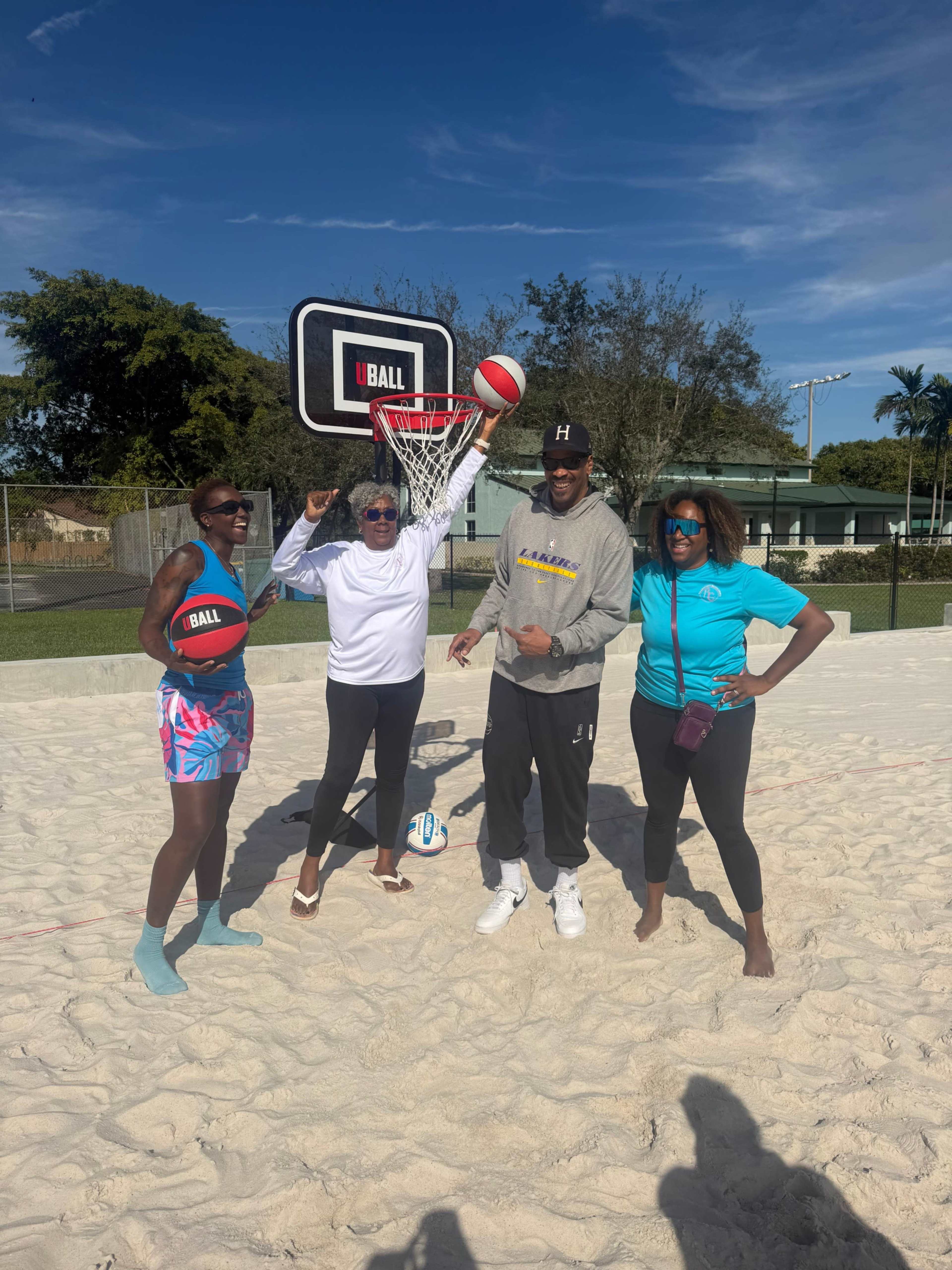 Four individuals pose on a sand court near a basketball hoop, holding balls and smiling.
