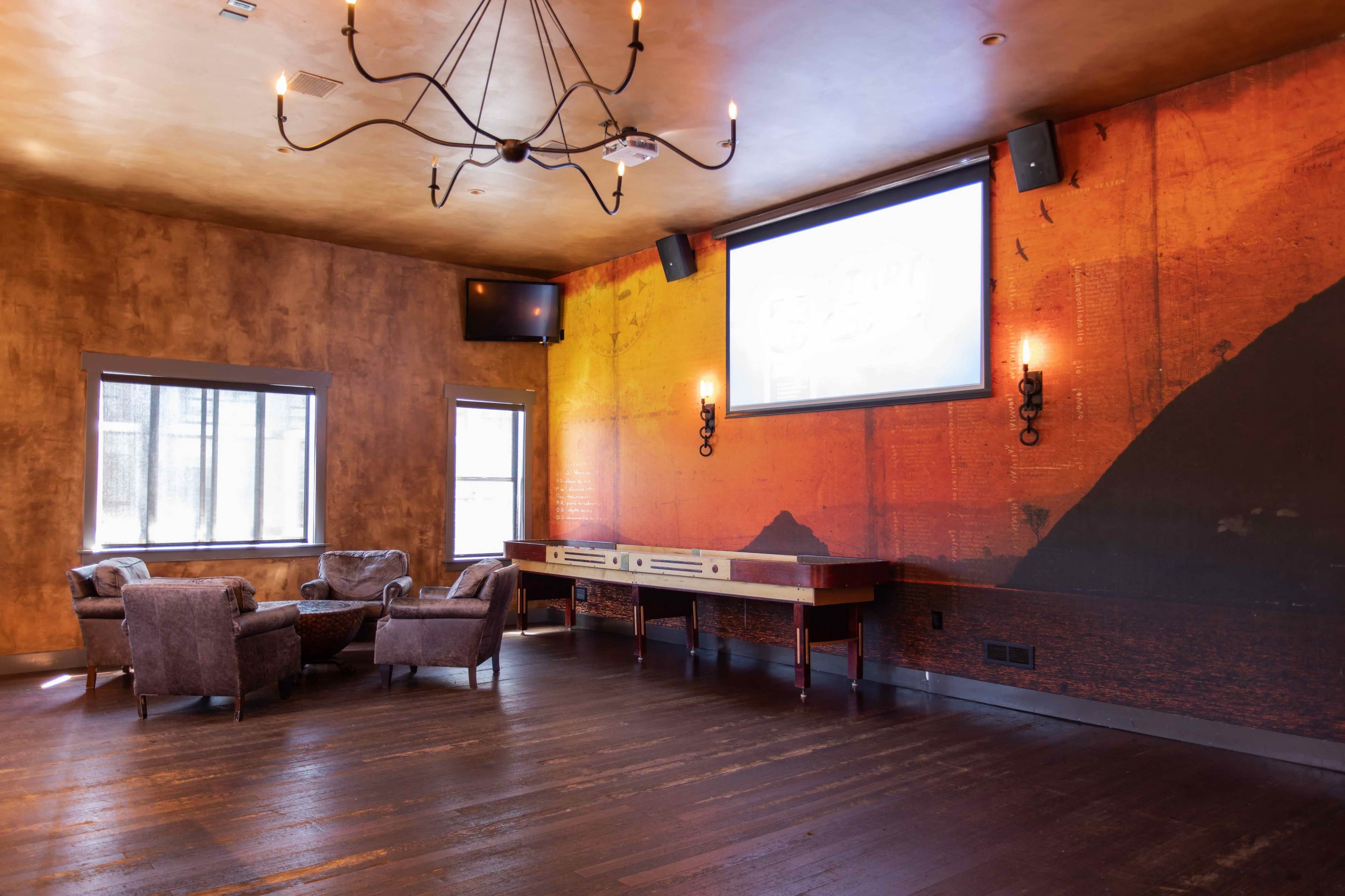 A modern lounge area with two plush armchairs facing a wooden console beneath a large screen, set against a textured wall featuring an orange and black mural.