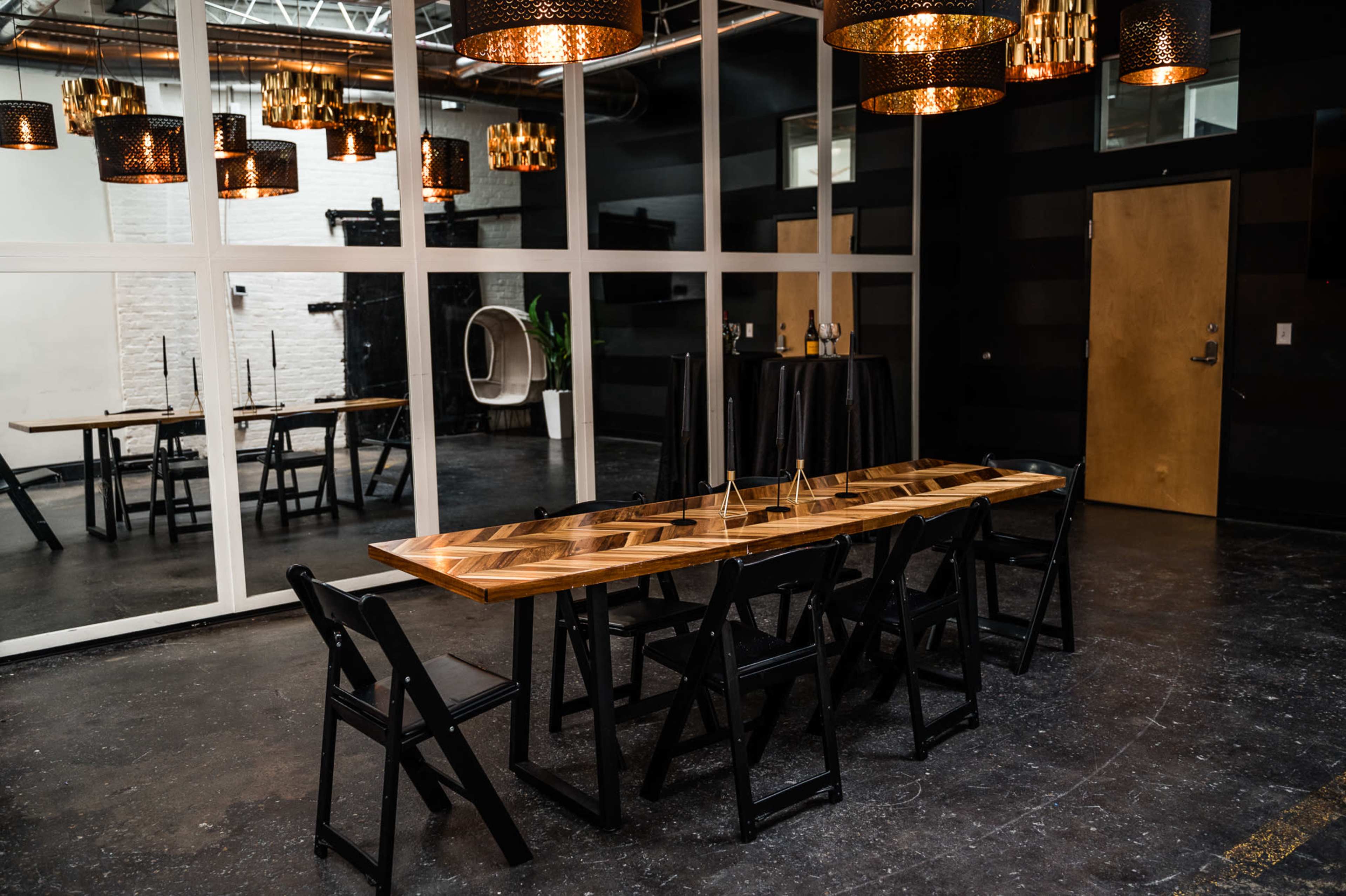 A long, rectangular wooden table with black folding chairs is set in a spacious room featuring large windows, modern lighting fixtures, and mirrored walls.